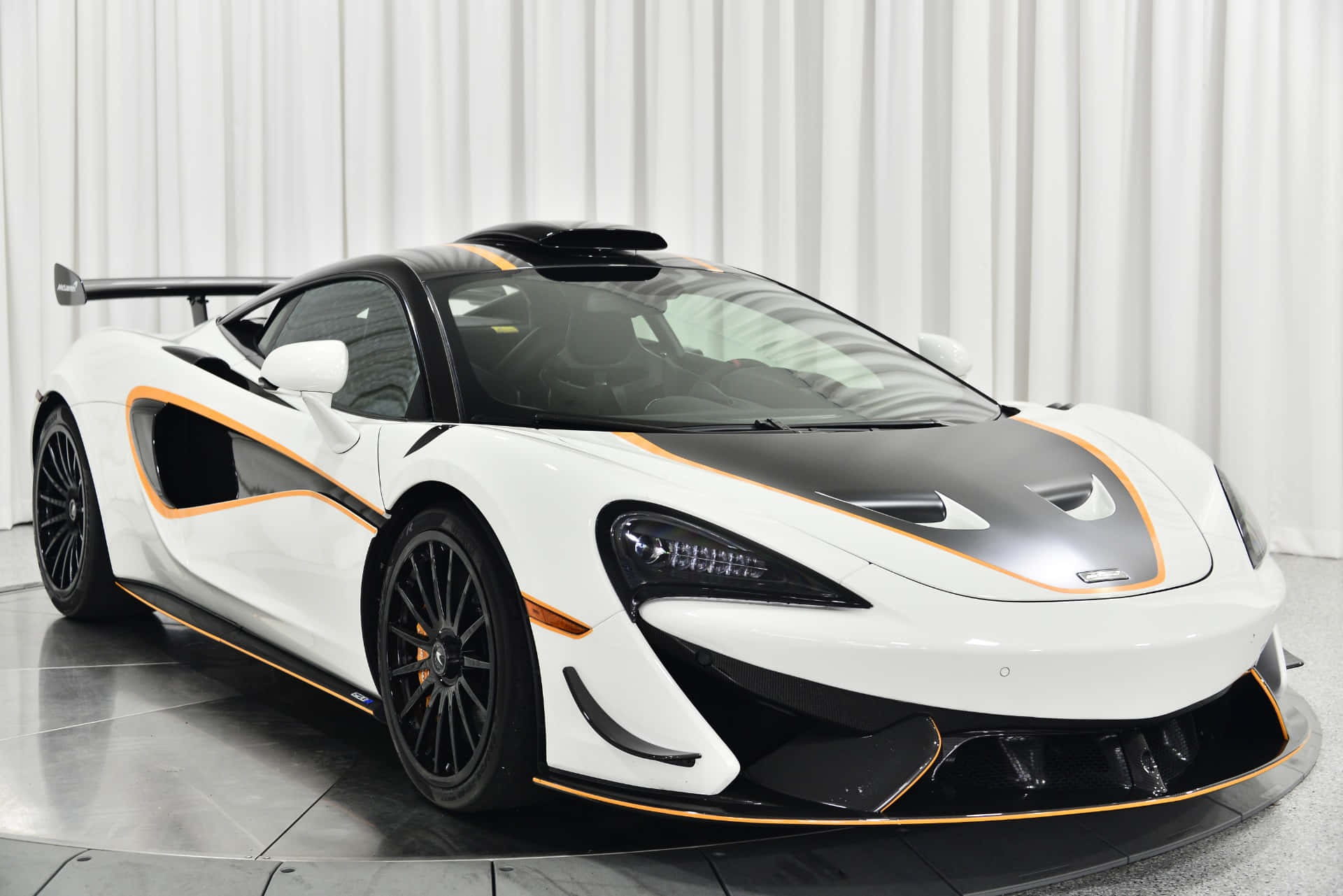 Mclaren 620r 桌布 壁紙 — 一輛時尚的白色McLaren跑車，帶有黑色和橙色點綴，在白色窗簾背景前的旋轉平台上展示。