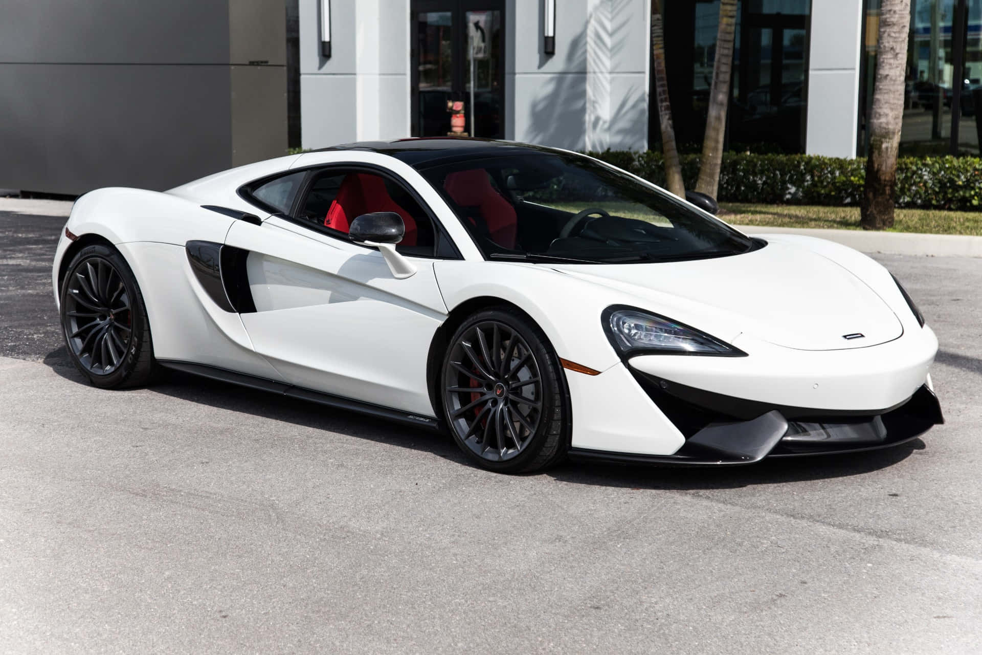 Mclaren Gt 桌布 壁紙 — 一架時尚的白色McLaren跑車，紅色內飾，停在有棕櫚樹的現代建築前的瀝青路上。