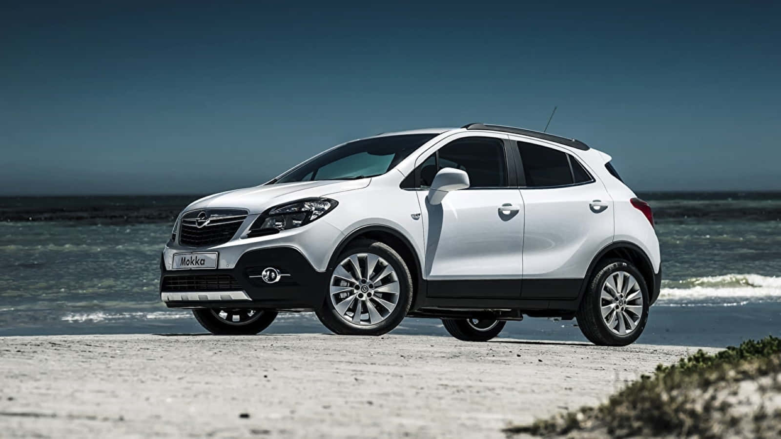 Opel Mokka 桌布 壁紙 — 一架時尚的白色Opel Mokka SUV停泊在沙灘上，背後有海浪拍打，天空清澈湛藍。