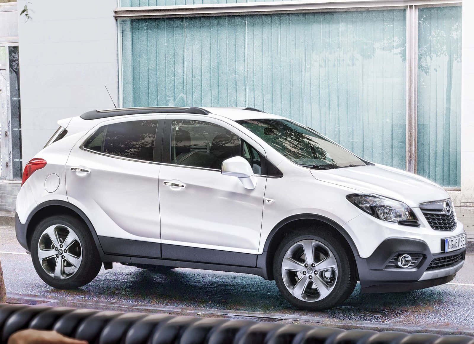 Opel Mokka 桌布 壁紙 — 一輛時尚的白色Opel Mokka SUV停泊在濕潤的城市街道上，旁邊是一座帶有垂直百葉窗的現代建築。