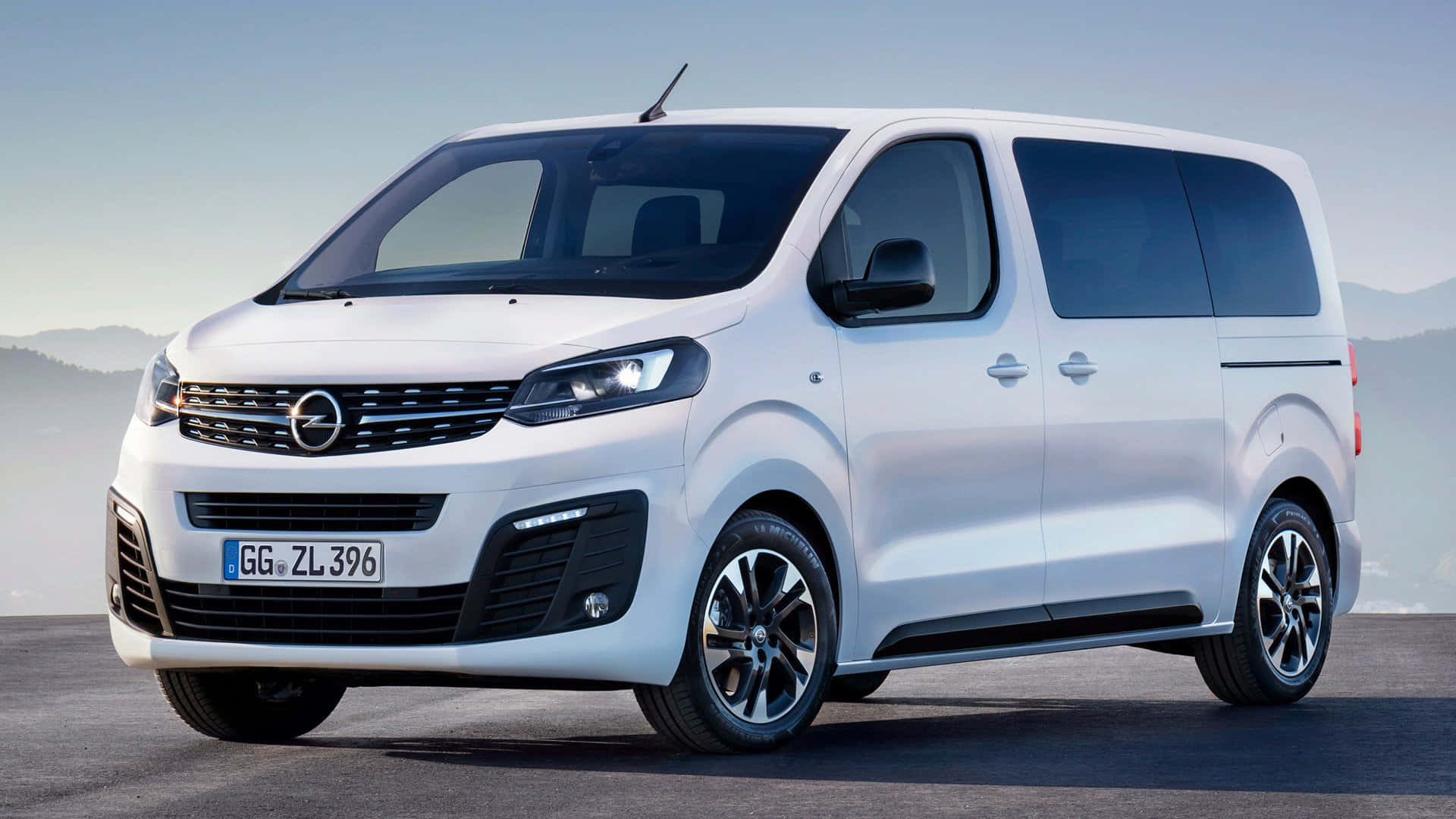 Opel Zafira 桌布 壁紙 — 一架時尚的白色Opel Vivaro客貨車停泊在鋪砌路面上，背景是朦朧的山巒，天空晴朗。