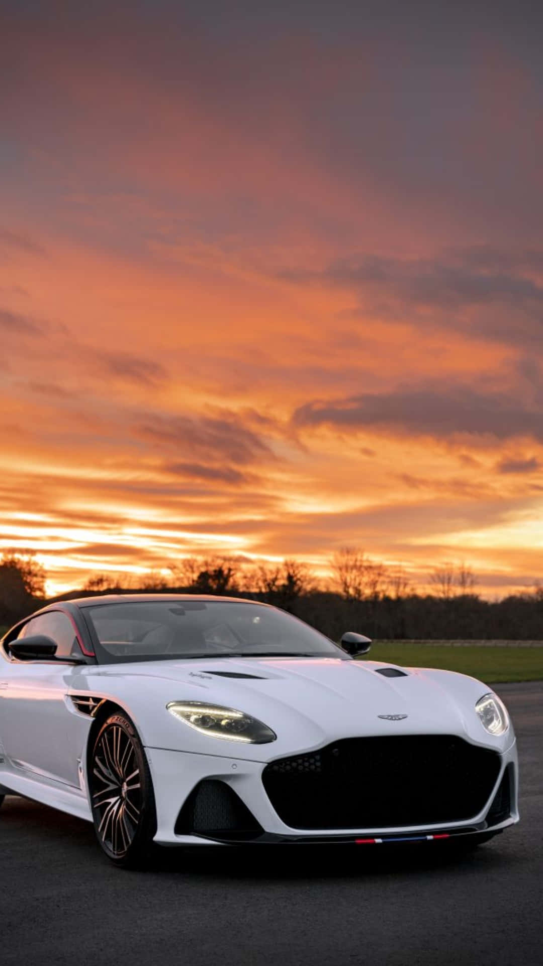 Aston Martin Dbs Superleggera 桌布 壁紙 — 一輛時尚的白色跑車在火紅橙色和粉紅色的日落天空下閃閃發光，伴隨著烏雲。