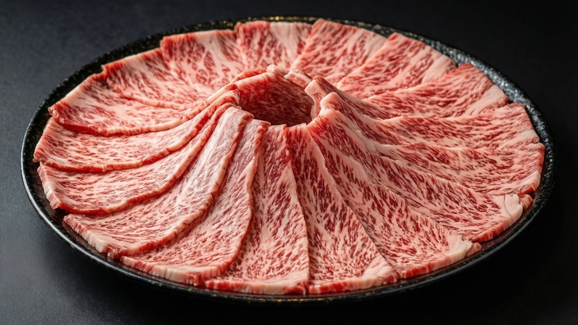 Premium Kobe Beef for Shabu 壁紙 — 在黑色背景的深色斑點盤子上，以扇形排列的大理石紋和牛切片。