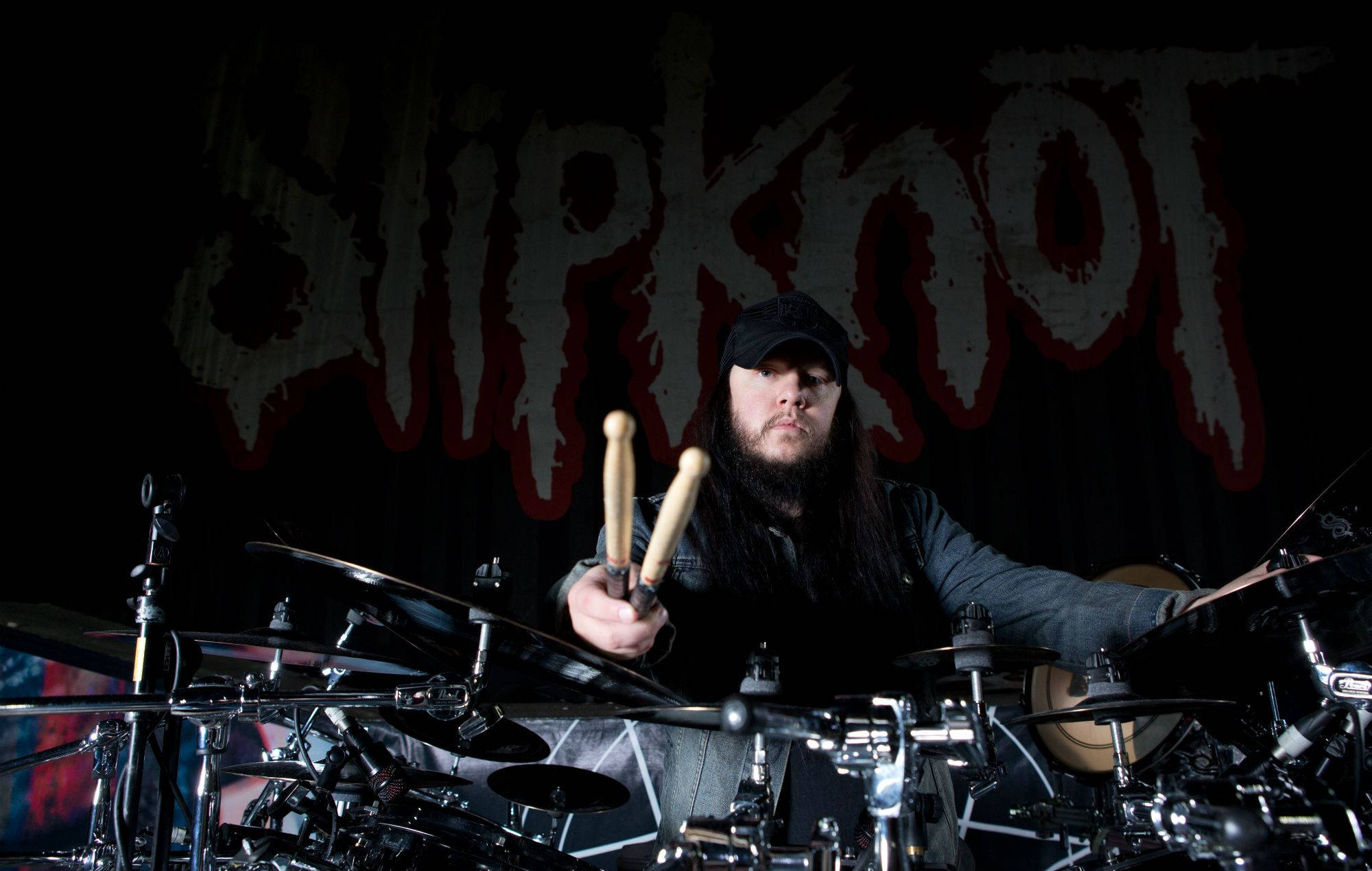 喬伊·喬迪森Slipknot鼓手 壁紙 — 一位留鬍鬚的鼓手戴着黑色帽子，在戲劇性的舞台燈光下，於金屬鼓組前手持鼓棍，身後是血紅色的Slipknot標誌。