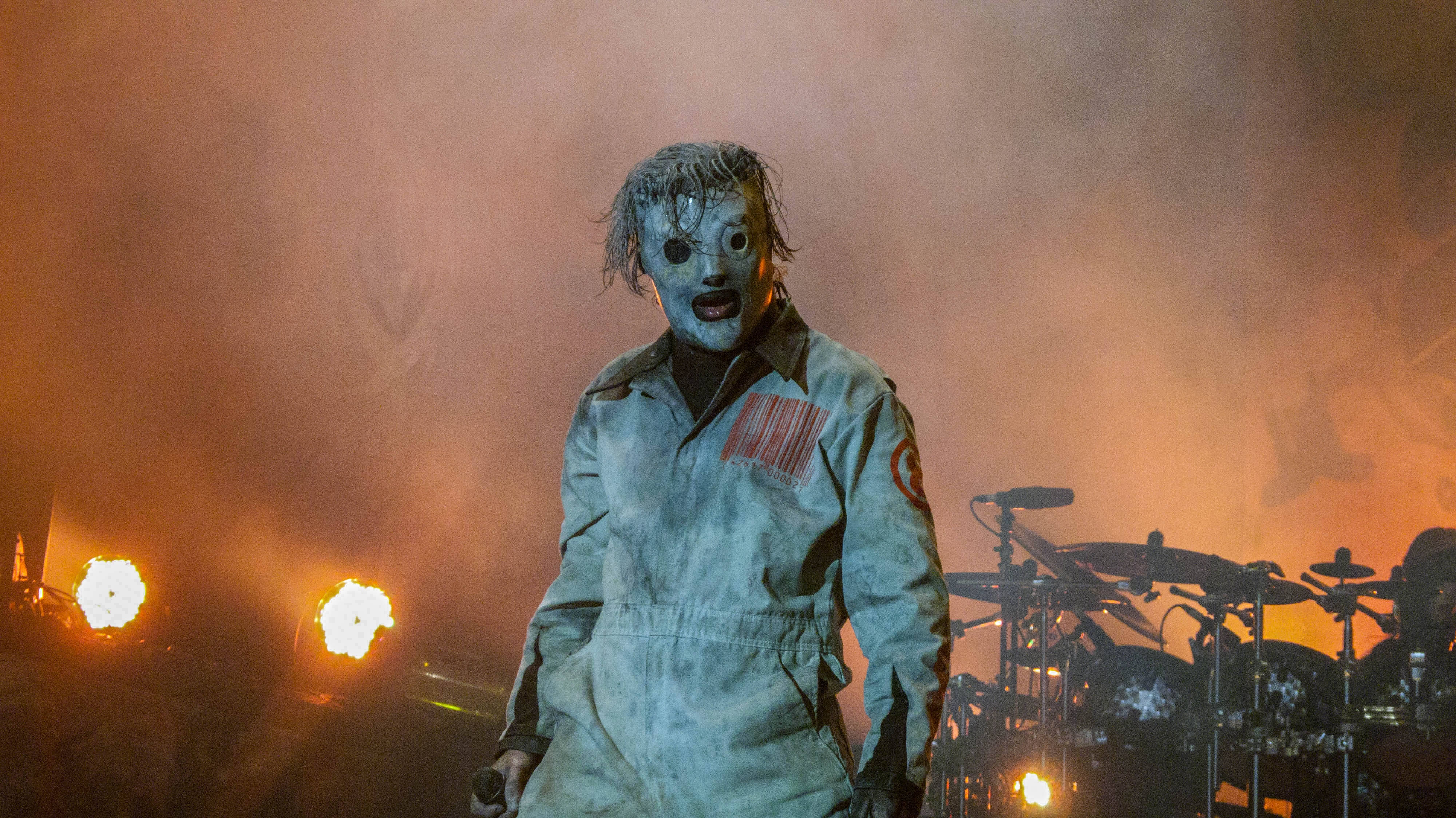 Slipknot Sid熱舞舞台 壁紙 — 一位戴著面具的表演者穿著骯髒的連身衣，在煙霧彌漫的舞台上，沐浴在火熱的橙色燈光中，熱情地歌唱。