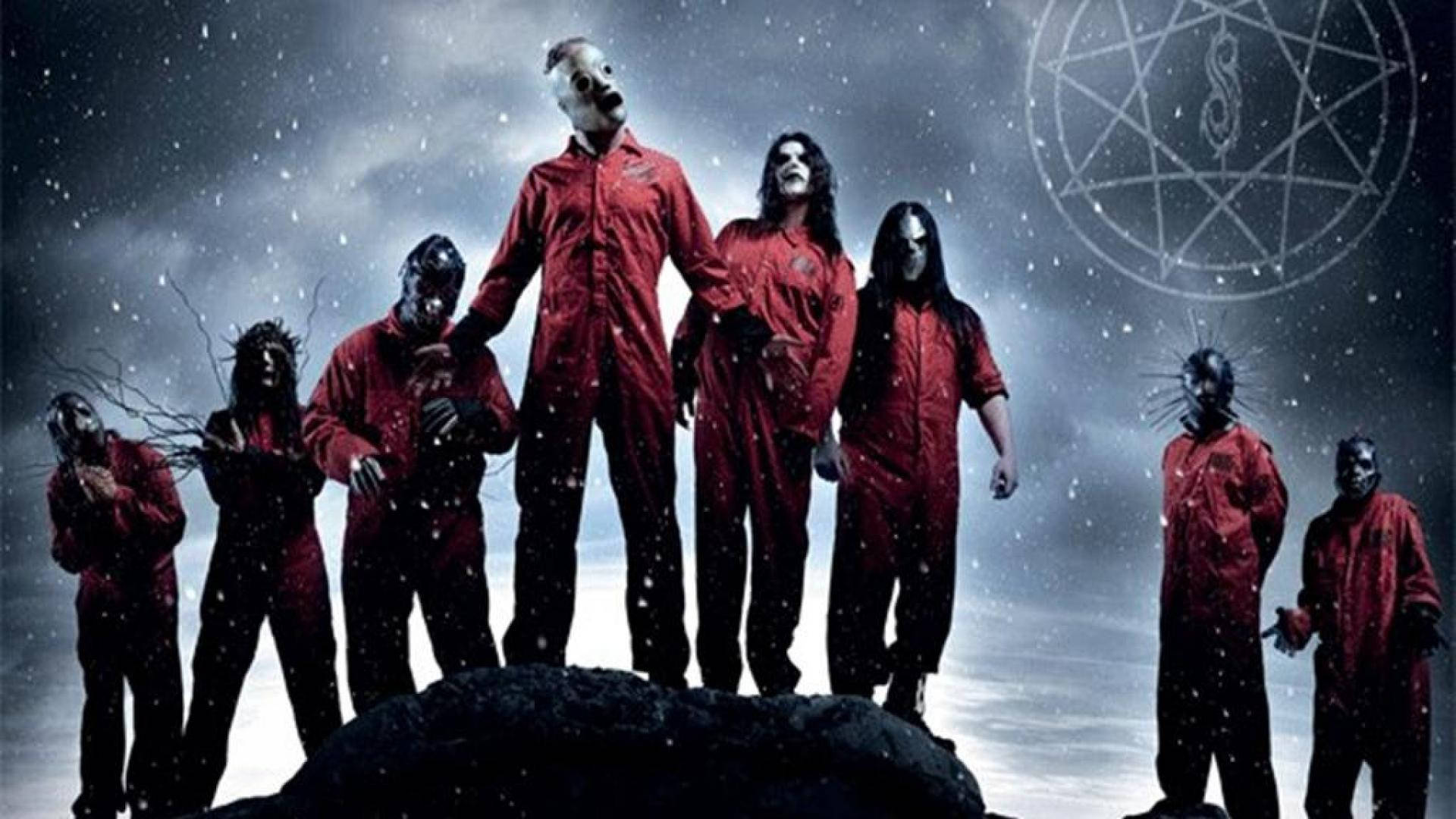 Slipknot 成員穿囚服壁紙 壁紙 — 七個戴面具、身穿紅色連身衣的人物站在雪覆蓋的岩石上，暴風雨的天空下，上方有一個發光的五角星。