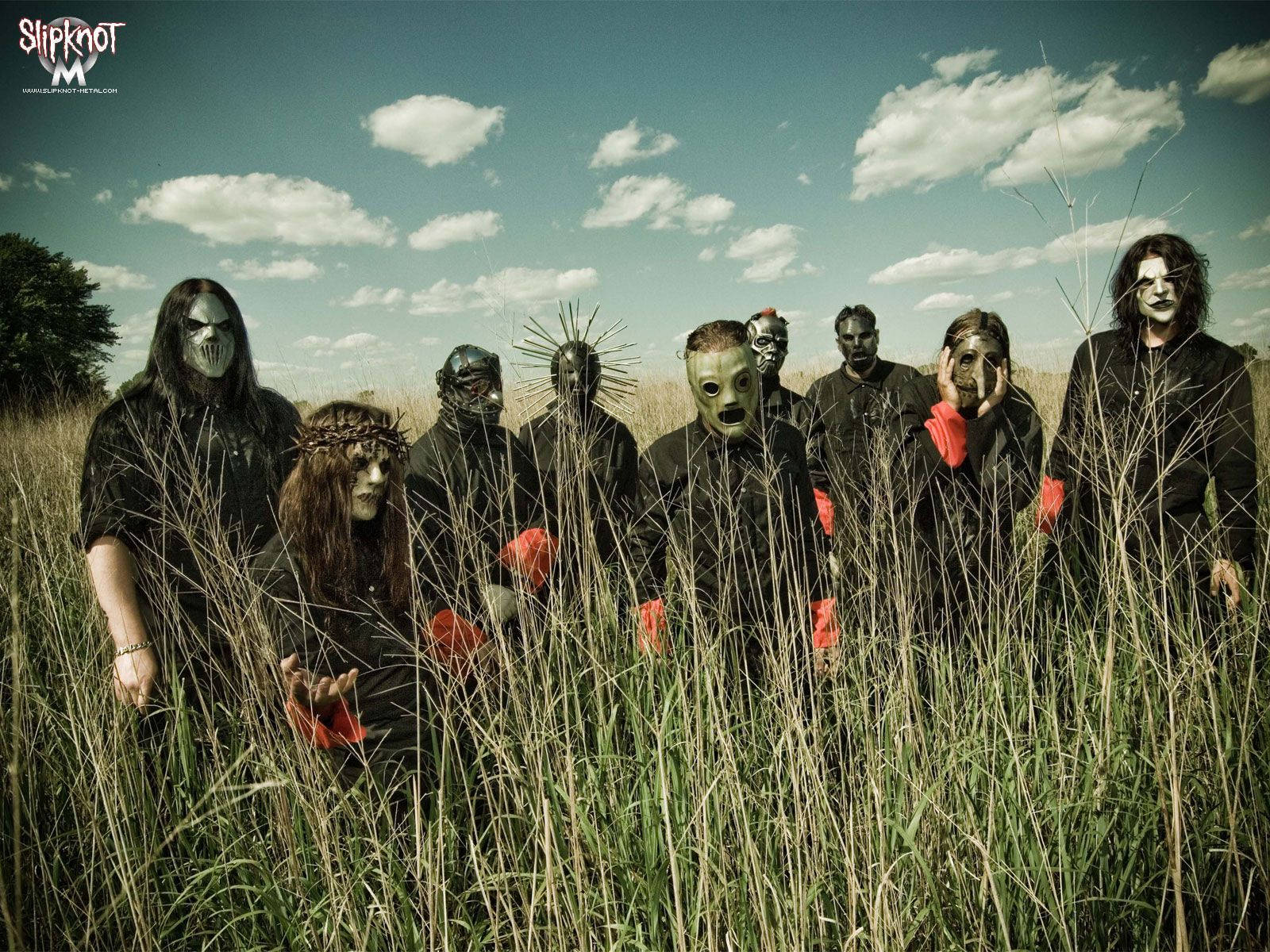 Slipknot 成員立於青草之上 壁紙 — 一個戴著面具、身穿黑色服裝的樂隊站在高高的草叢中，天空多雲。