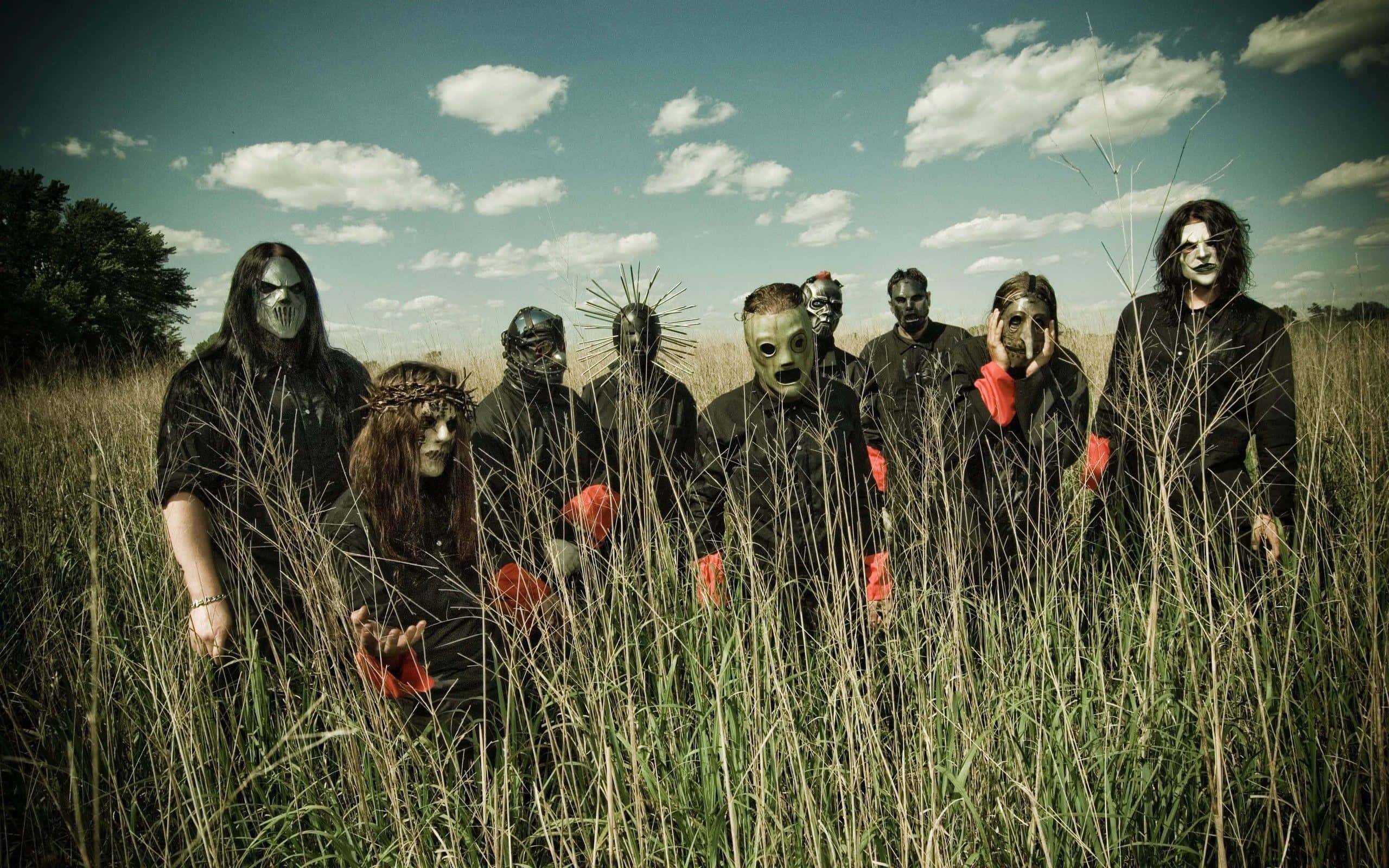 Slipknot 桌面男團在田野中 壁紙 — 一群戴著面具、身穿黑色衣服和紅色手套的人站在多雲天空下的高草叢中。