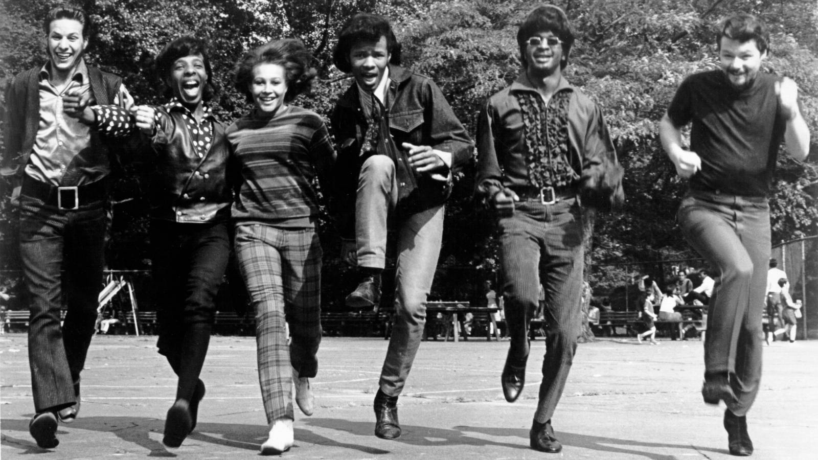 Sly 及 The Family Stone 黑白照片 壁紙 — 六個穿著1970年代時尚的人，在陽光普照的公園廣場上充滿活力地跳躍，背景有樹木和長椅。