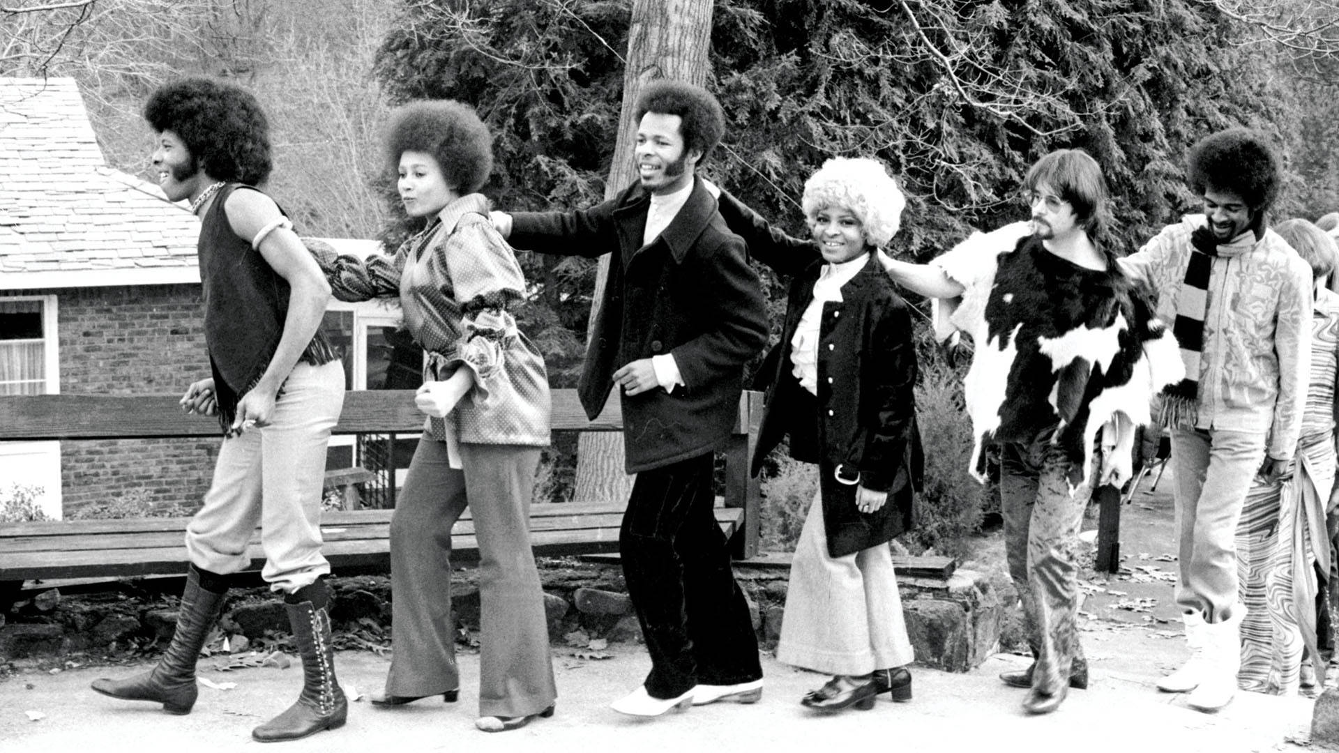 Sly與Family Stone的搞笑合照 壁紙 — 一張黑白照片，六個身穿1970年代時尚的人士在戶外擺姿勢，背景有樹木和房屋。