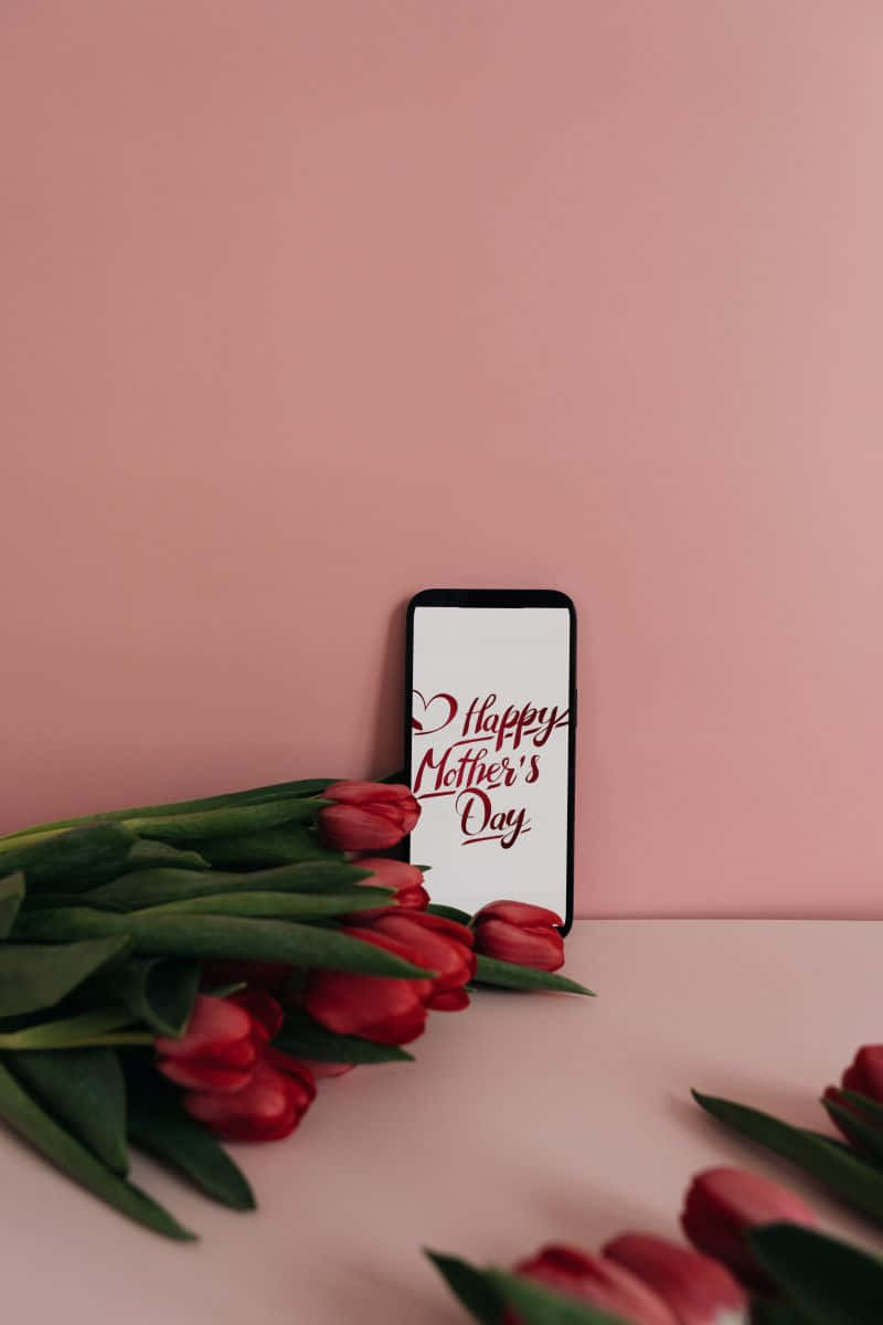  壁紙 — 一部智能手機顯示紅色字體的「Happy Mother's Day」，旁邊放著一束紅色鬱金香，背景是柔和的粉紅色。