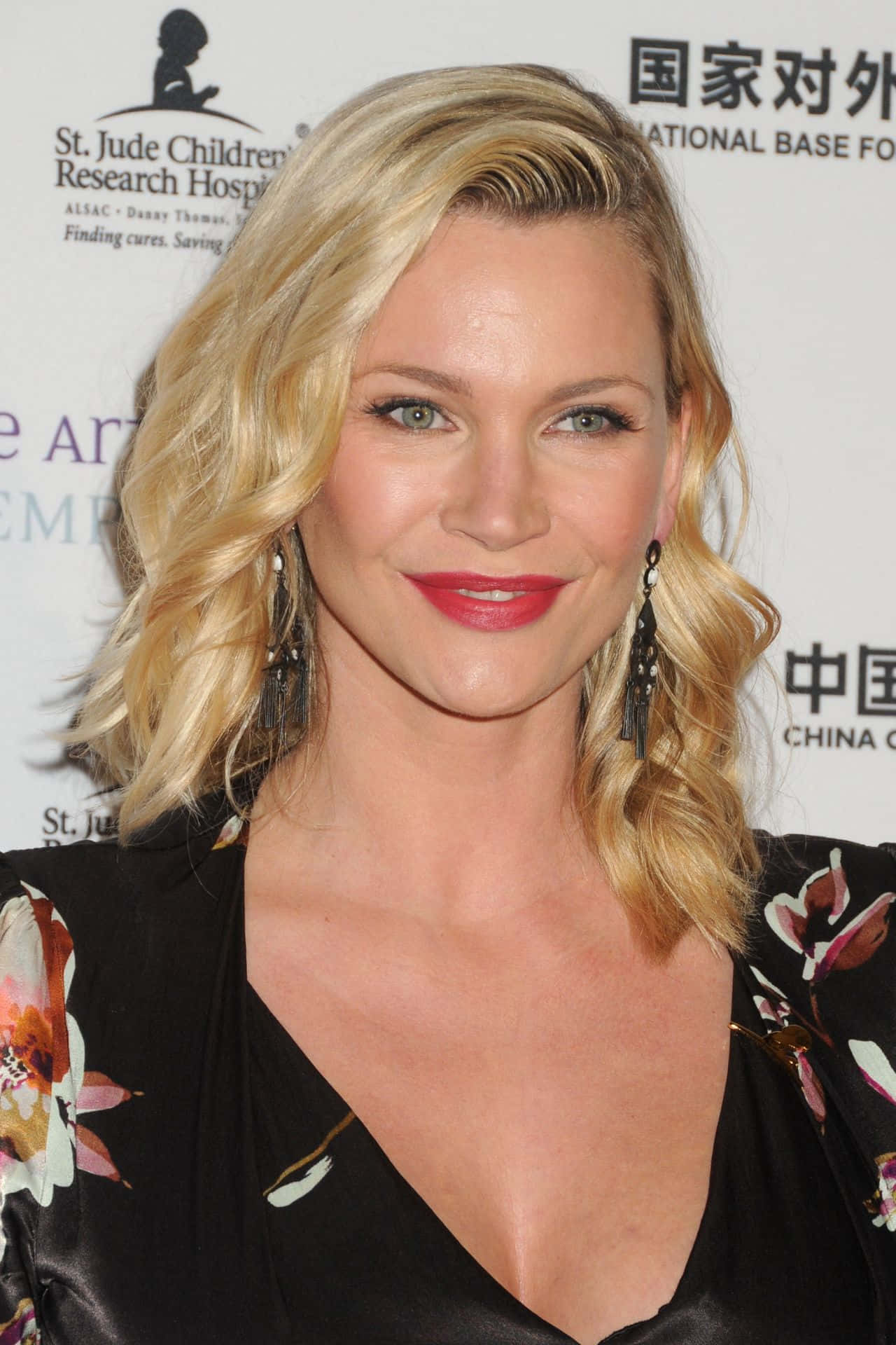 Natasha Henstridge 桌布 壁紙 — 一位微笑的金髮女性，塗著紅色唇膏，穿著花卉襯衫，在品牌背景前於慈善活動中擺姿勢。