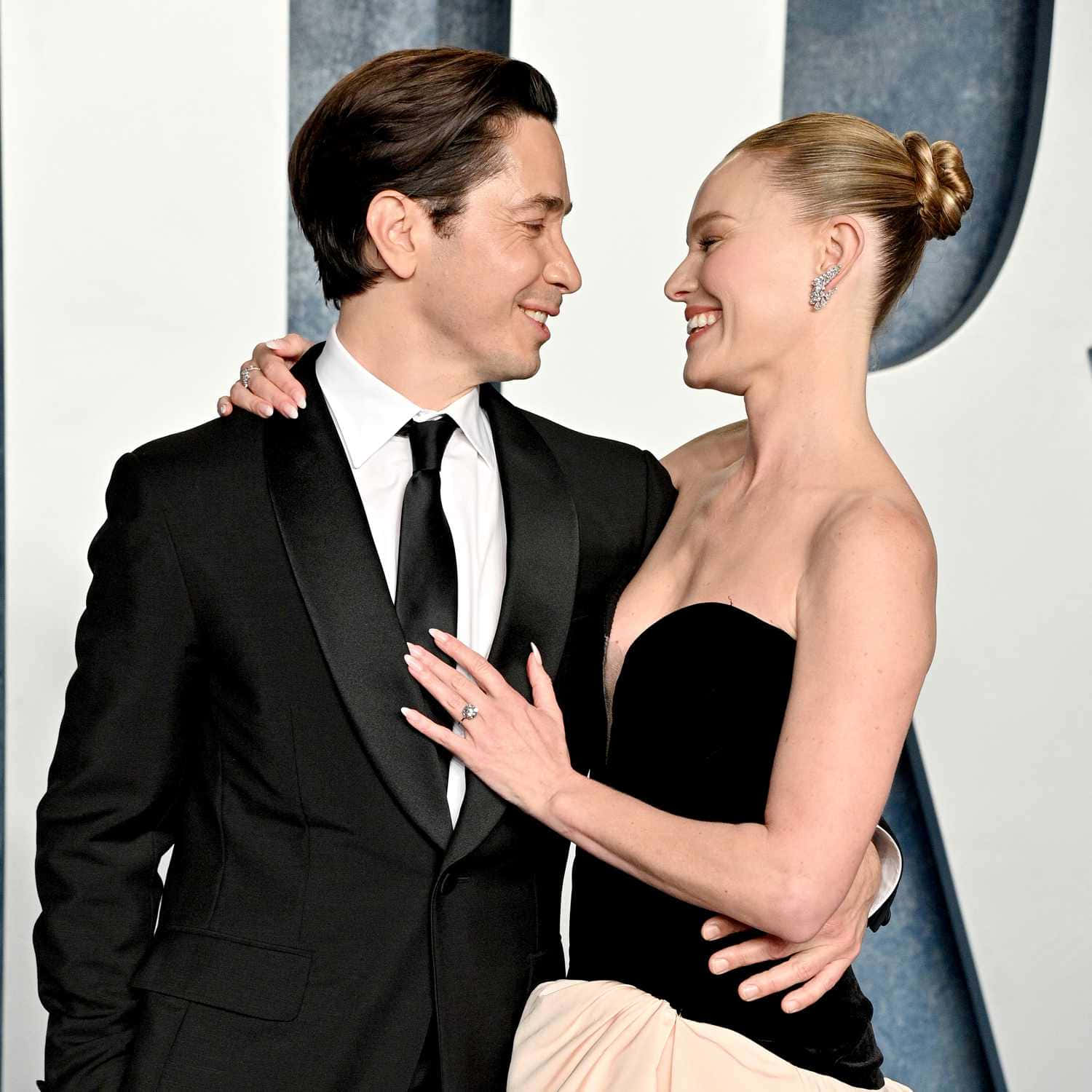 Kate Bosworth 桌布 壁紙 — 一對微笑的情侶穿著正式黑色服裝，在時尚的藍白背景下溫暖擁抱。