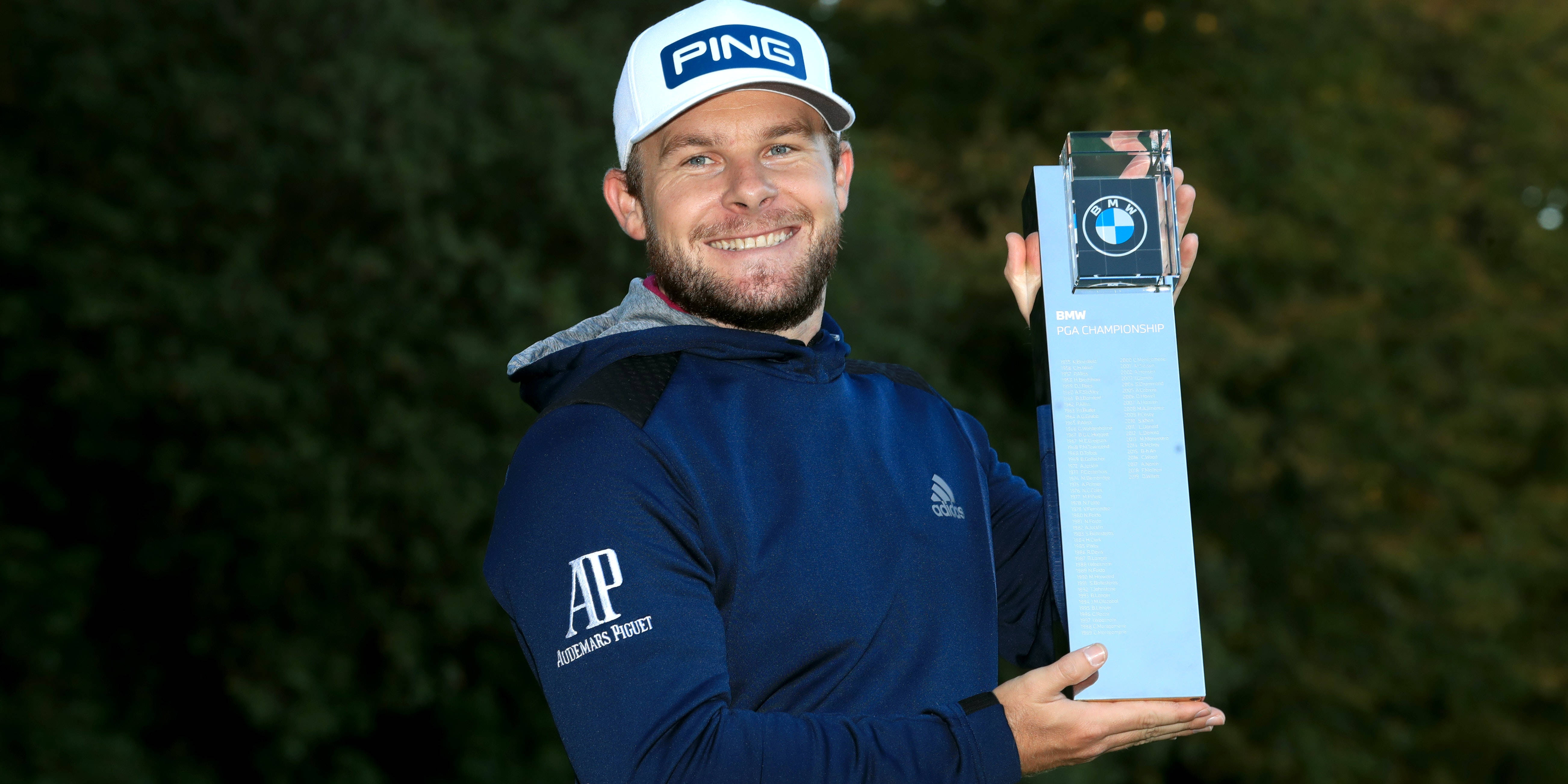 Tyrrell Hatton 桌布 壁紙 — 一位面帶微笑的高爾夫球手，身穿藍色連帽衫和白色帽子，在模糊的綠色背景下自豪地高舉BMW PGA錦標賽獎盃。
