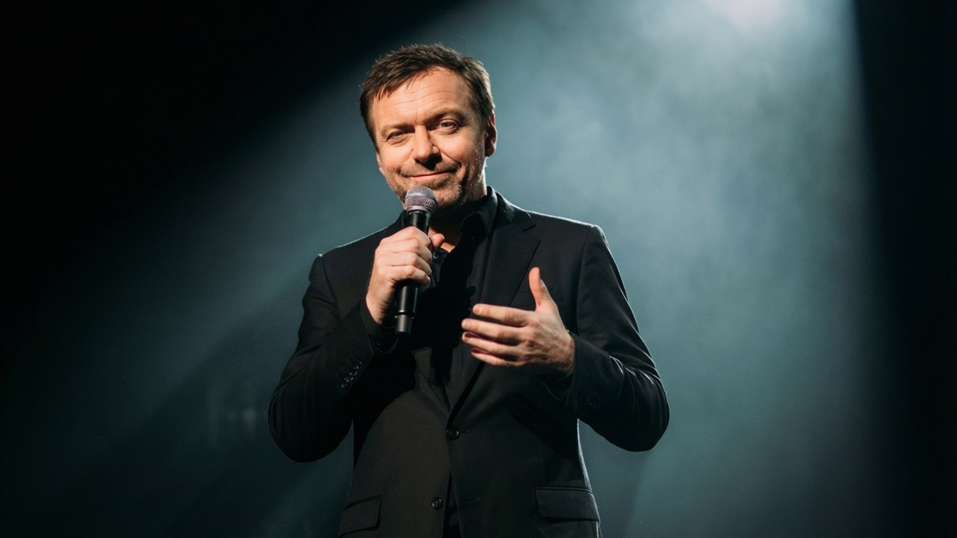 ricky gervais 桌布 壁紙 — 一位微笑的黑西裝男子在聚光燈下，於昏暗的舞台上手持麥克風，周圍瀰漫著氣氛薄霧。