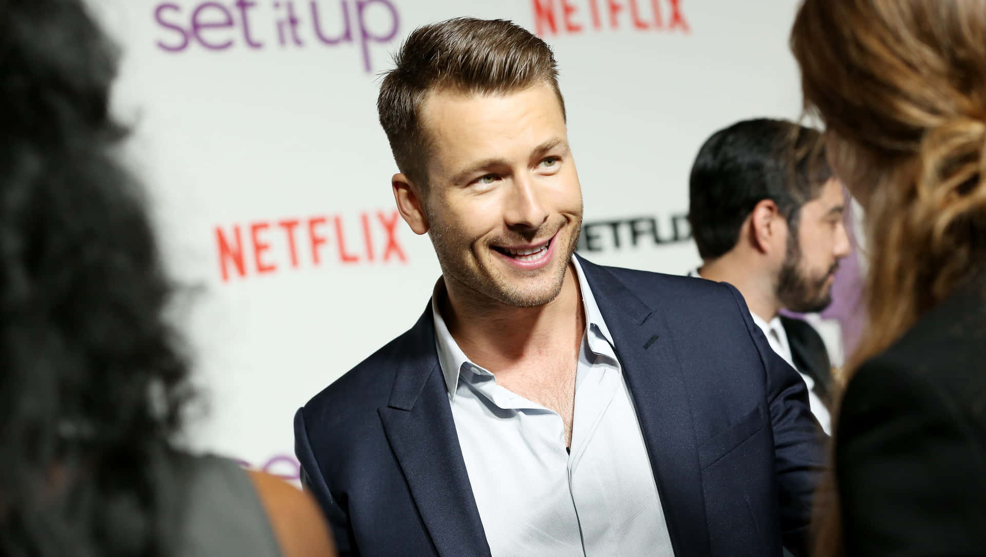  壁紙 — 一位微笑的男子身穿海軍藍西裝外套和淺色襯衫，在Netflix活動中，背後有「set it up」品牌標誌。