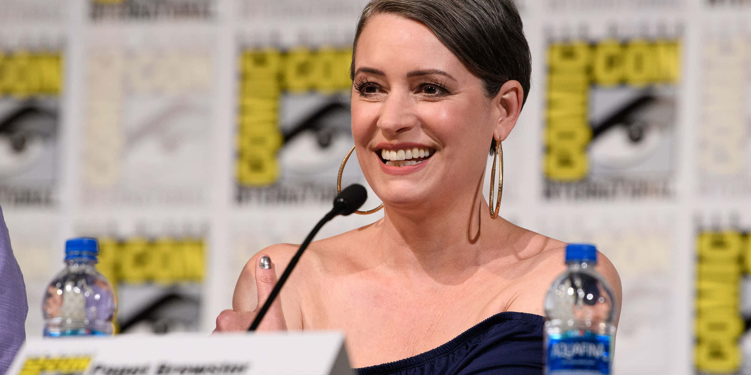 paget brewster 桌布 壁紙 — 一位微笑的短黑髮女性戴著金色耳環，在帶有品牌背景和水樽的Comic-Con座談會上發言。