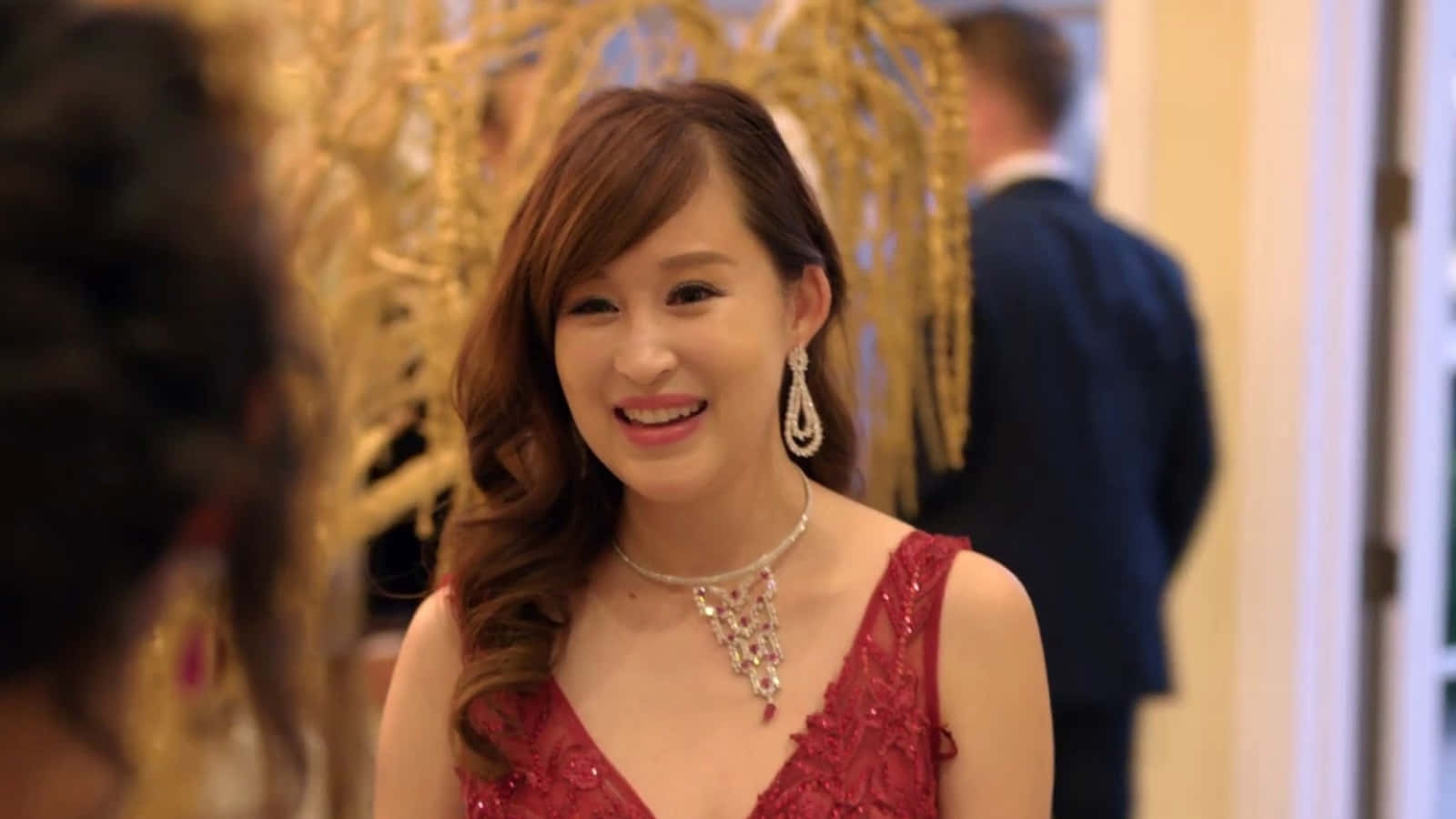 Bling Empire 桌布 壁紙 — 一位微笑的女性，身穿紅色珠飾連衣裙和閃閃發光的珠寶，站在一個華麗的金色裝飾房間裡。