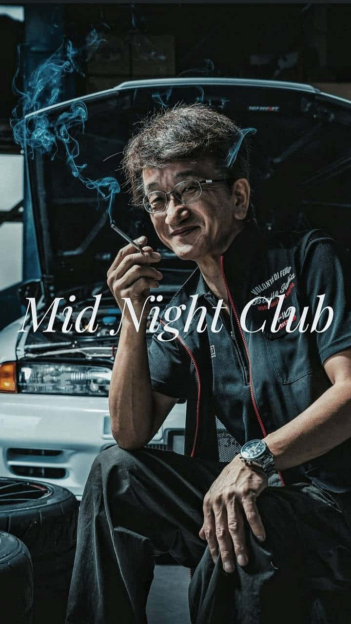 日本汽車工程師Smokey Nagata 壁紙 — 一個戴眼鏡的男人坐在一輛引擎蓋打開的汽車前，在燈光昏暗的車庫裡抽煙。