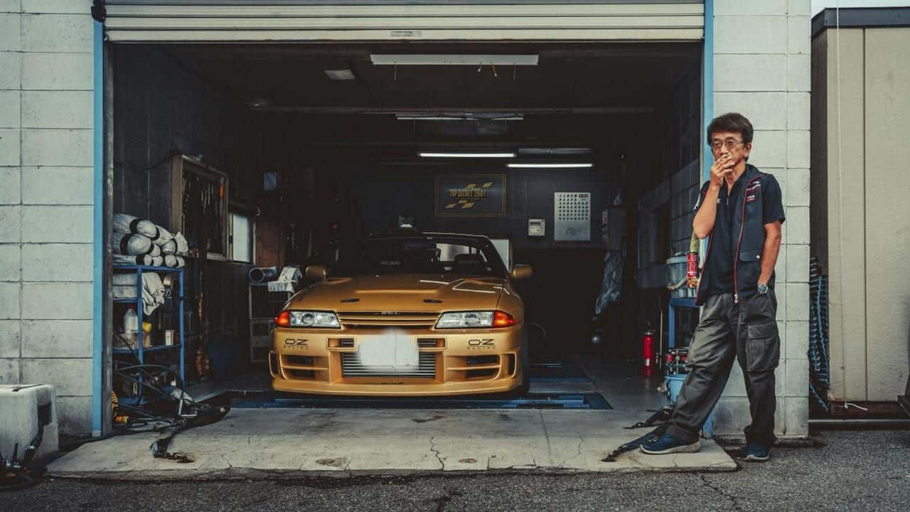 Smokey Nagata與金色跑車 壁紙 — 車房裡的機械師站在金色日產Skyline R32旁邊，周圍有工具和設備。