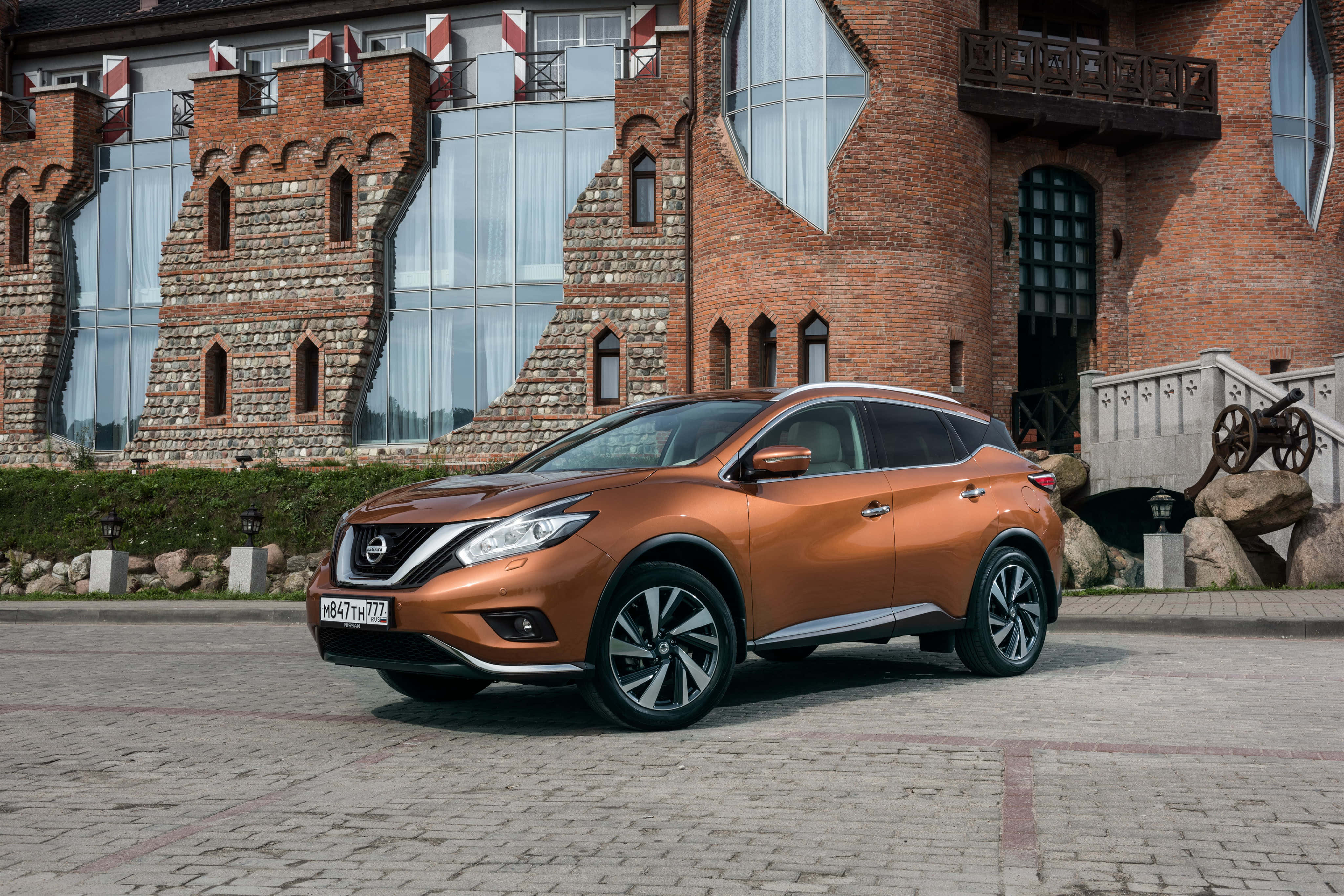Nissan Murano 桌布 壁紙 — 一輛時尚的銅色日產Rogue SUV停泊在石頭廣場上，前面是現代城堡風格的磚砌建築，配有玻璃窗。