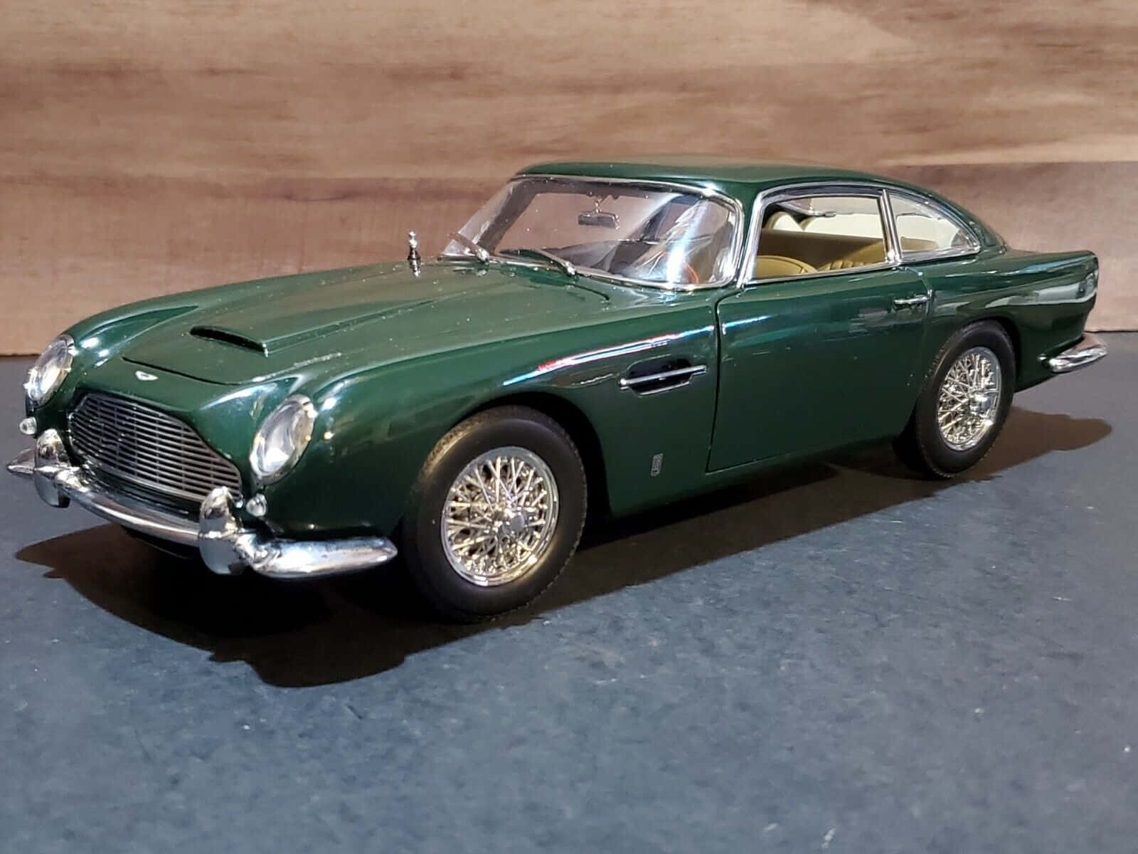 Aston Martin Db5 桌布 壁紙 — 一架光滑的綠色Aston Martin DB5模型車，帶有鍍鉻裝飾和鋼絲輪，放置在深色紋理表面上，背景為木質。