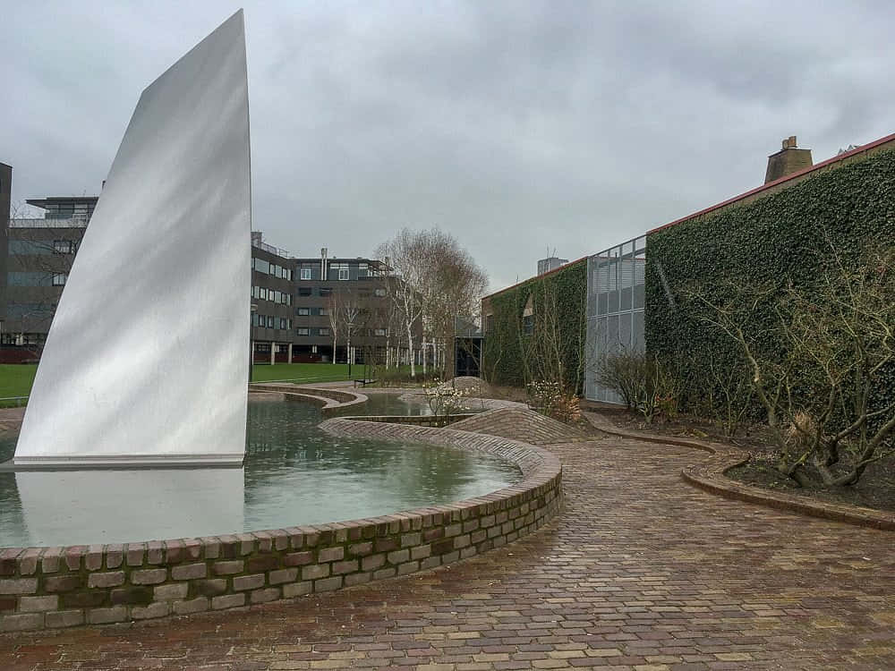 Tilburg 桌布 壁紙 — 一座光滑的白色帆形雕塑矗立在彎曲的磚砌池塘中，旁邊是一座被常春藤覆蓋的建築，天空多雲。