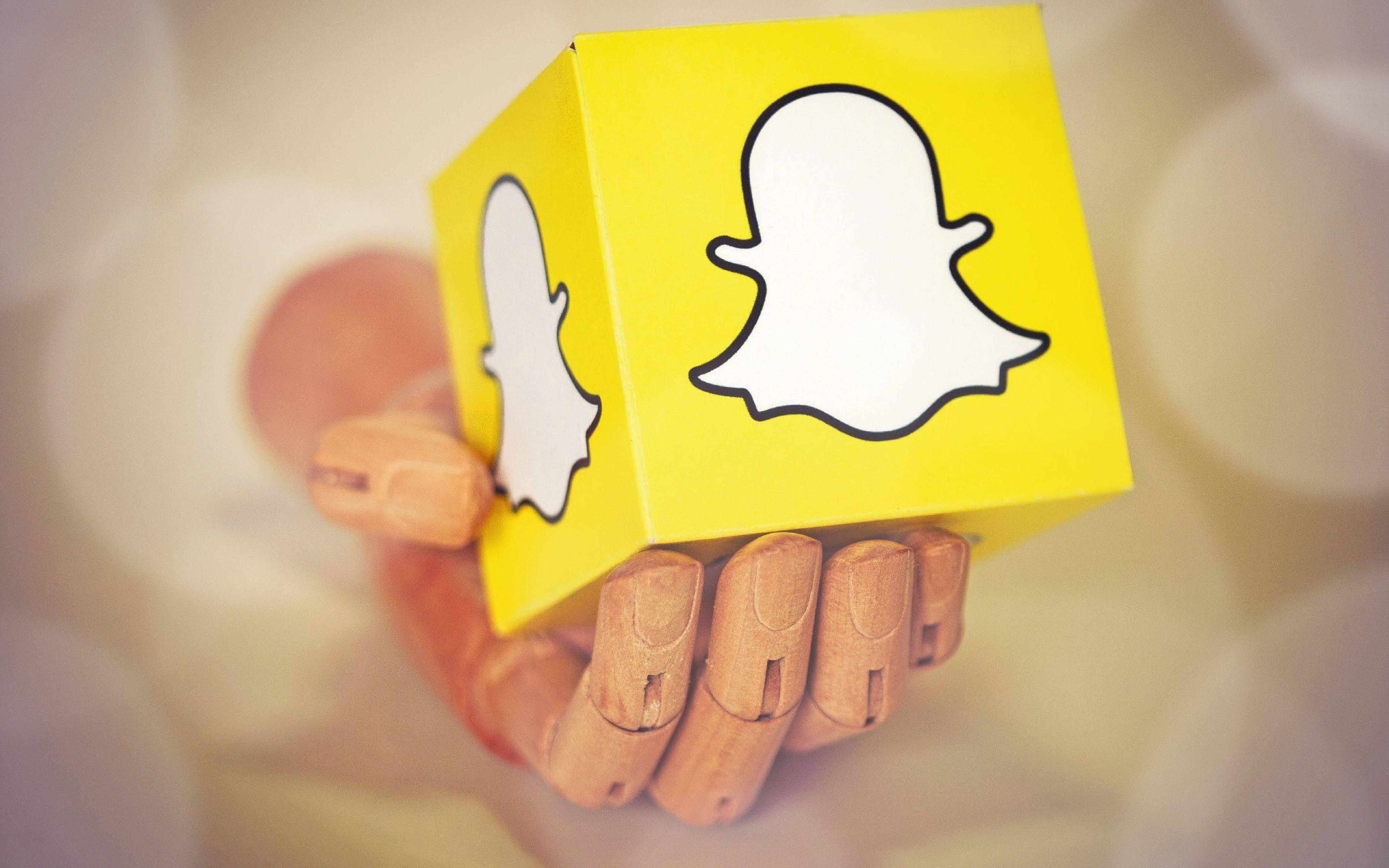 Snapchat標誌立方體3D藝術 壁紙 — 一隻木手拿著一個亮黃色立方體，上面有Snapchat鬼魂標誌，背景柔和模糊。