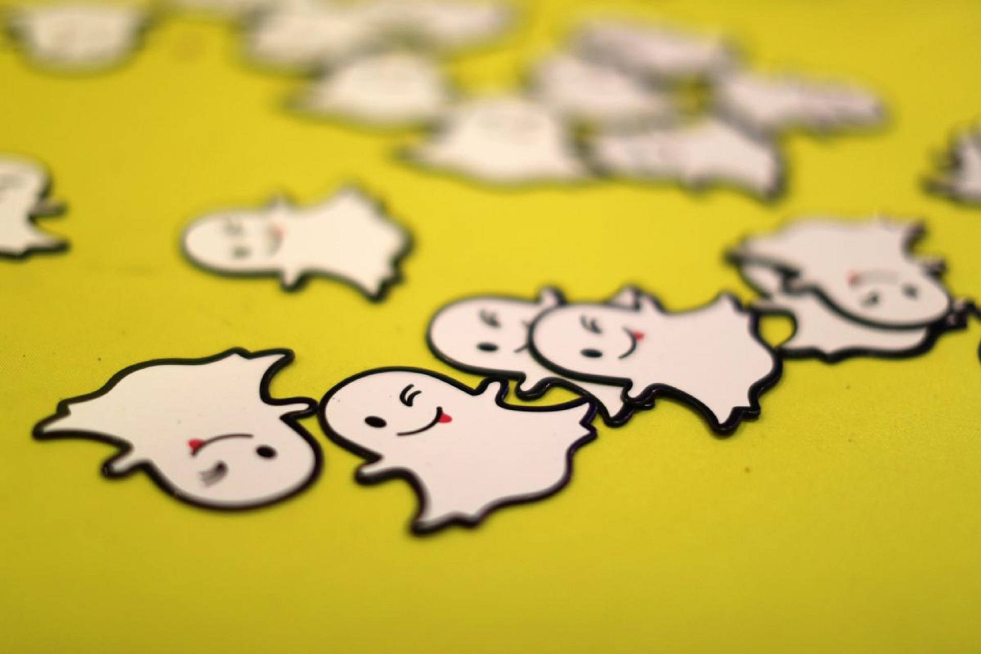 Snapchat標誌貼紙3D藝術 壁紙 — 淺景深突出了一組微笑的白色鬼魂形狀貼紙，帶有黑色輪廓，位於充滿活力的黃色表面上。
