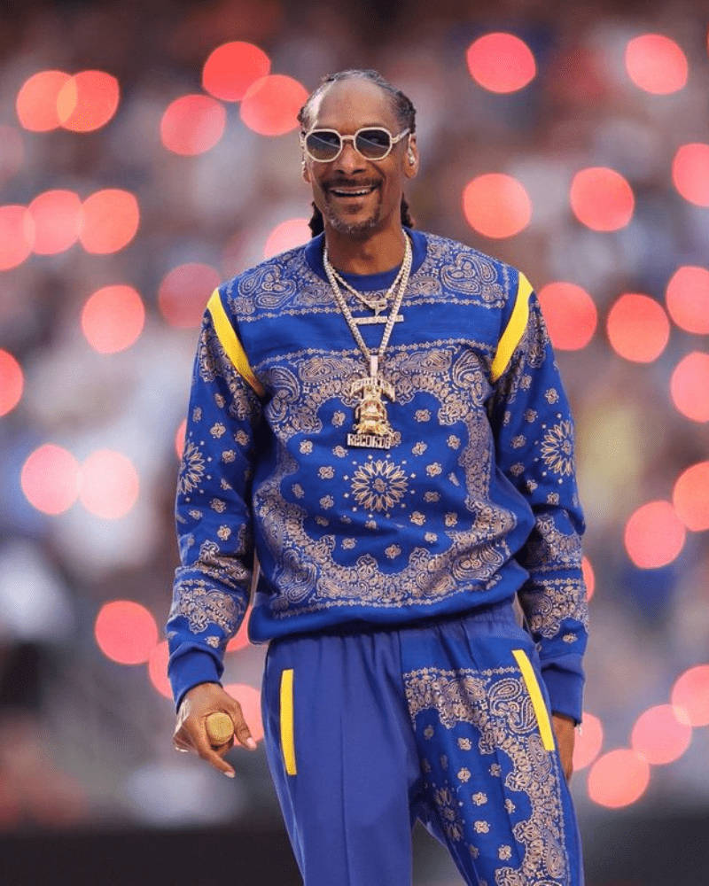 Snoop Dogg Crip 服裝 壁紙 — Snoop Dogg 自信地微笑，身穿藍色佩斯利花紋運動服，配金色點綴和一個大吊墜，背景是模糊的體育場和紅色散景燈光。