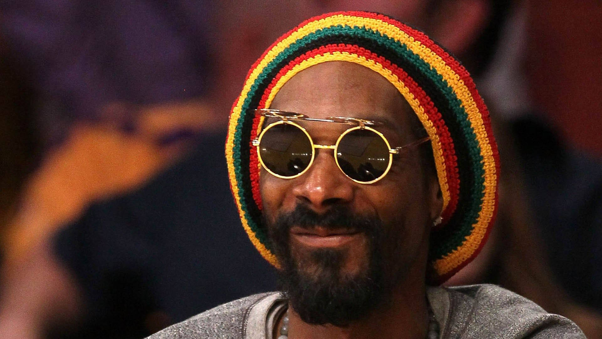 Snoop Dogg 戴拉斯特帽 壁紙 — Snoop Dogg 溫暖微笑，戴著圓形金色邊框太陽眼鏡和彩色條紋針織冷帽。