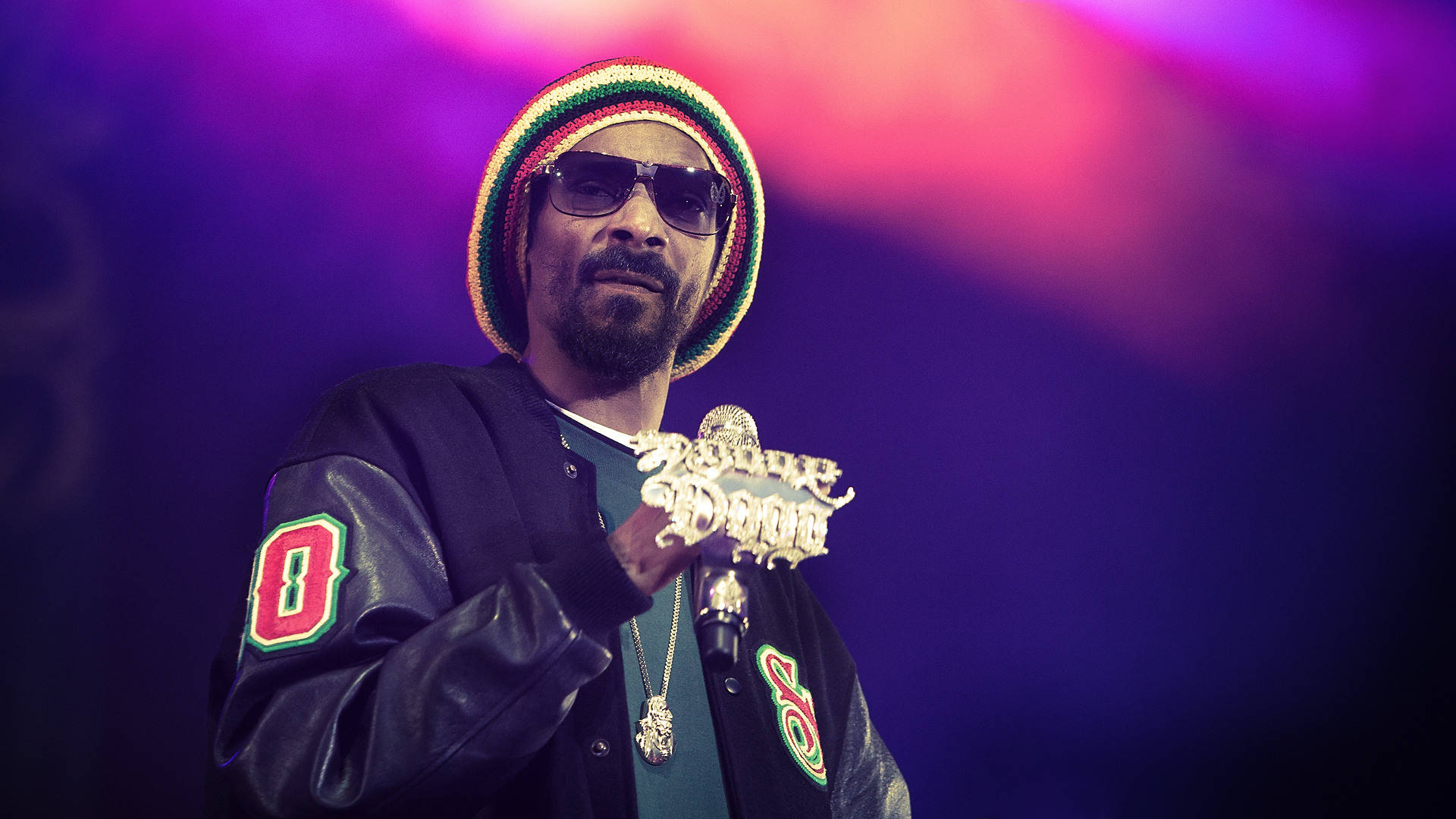 Snoop Dogg 持鑲嵌麥克風 壁紙 — Snoop Dogg 身穿黑色皮褸和彩色冷帽，在充滿活力的紫色和粉紅色舞台燈光下拿著金色麥克風。