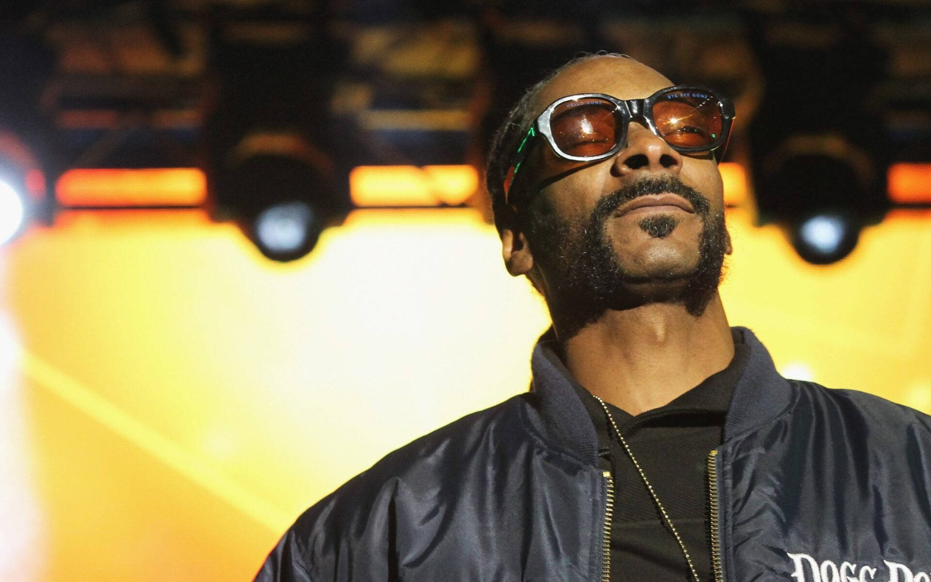 Snoop Dogg 演唱會現場 壁紙 — Snoop Dogg 戴著標誌性太陽眼鏡和深藍色飛行員夾克，在溫暖的舞台燈光下。