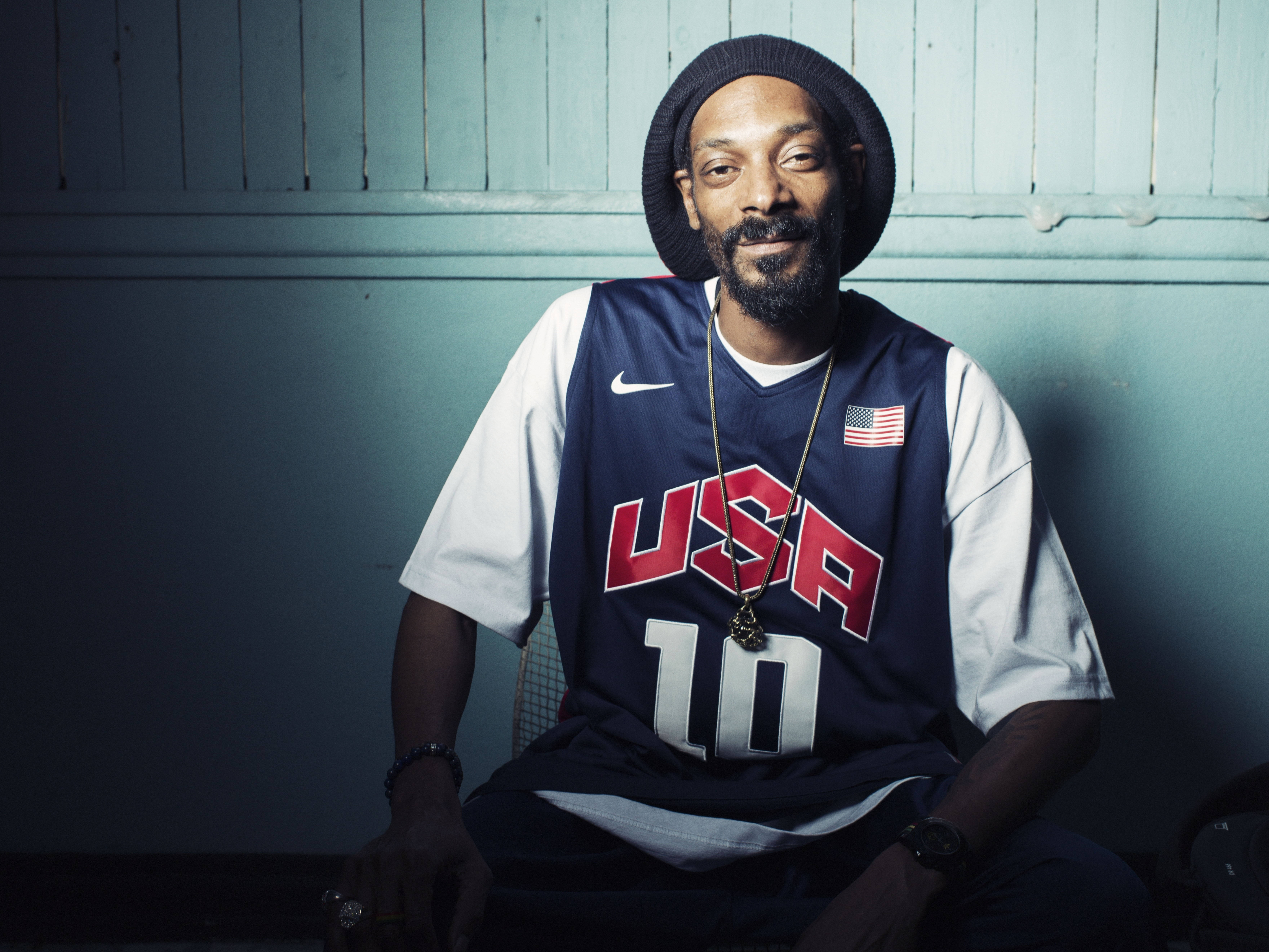 Snoop Dogg 穿著 Bryant 球衣 壁紙 — Snoop Dogg 隨意地坐在燈光昏暗的房間裡，身穿美國籃球球衣和冷帽，散發著酷酷的自信。