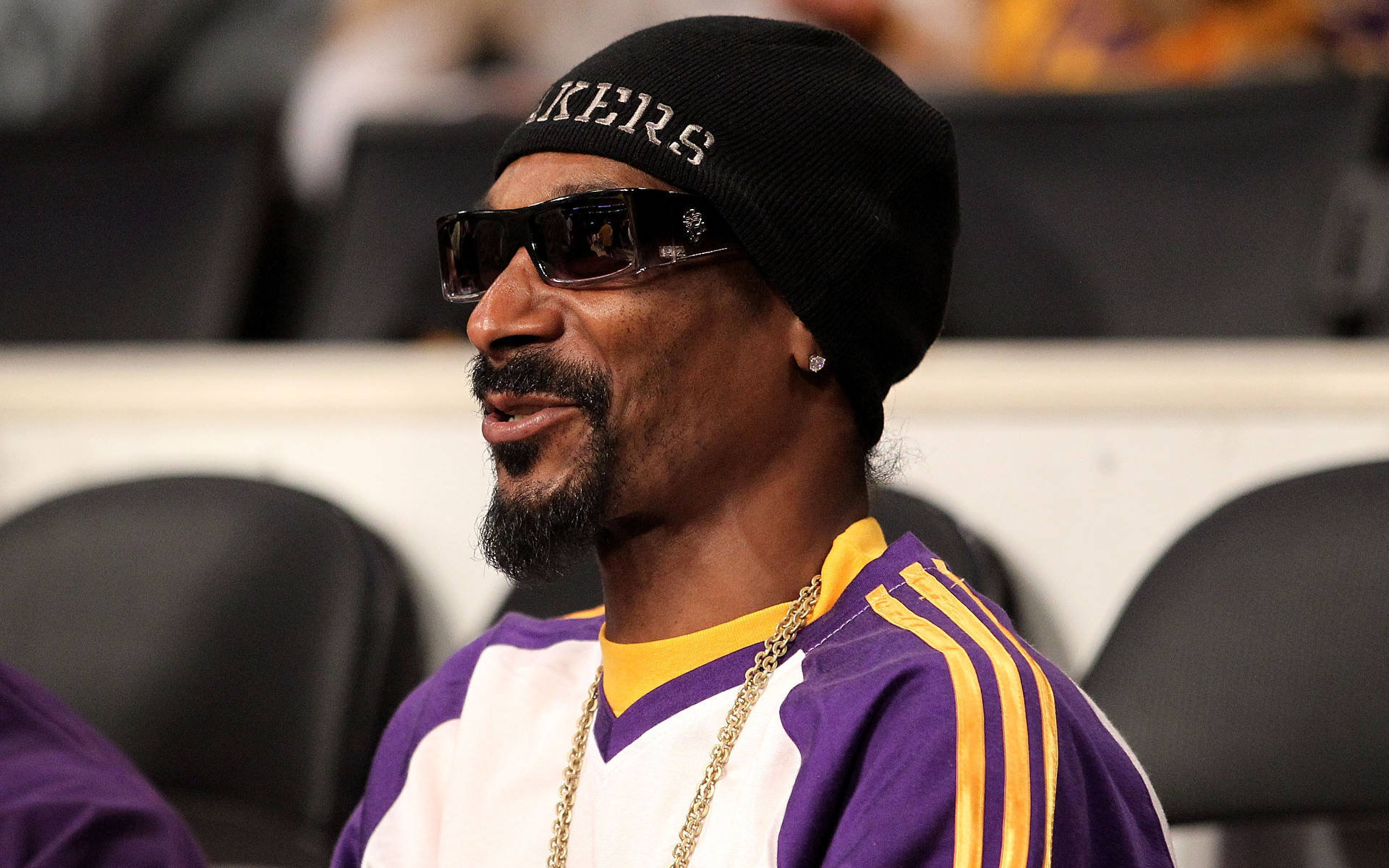 Snoop Dogg 觀看湖人隊比賽 壁紙 — Snoop Dogg 戴著黑色冷帽和太陽眼鏡，身穿紫色和金色的湖人球衣坐在體育館座位上。