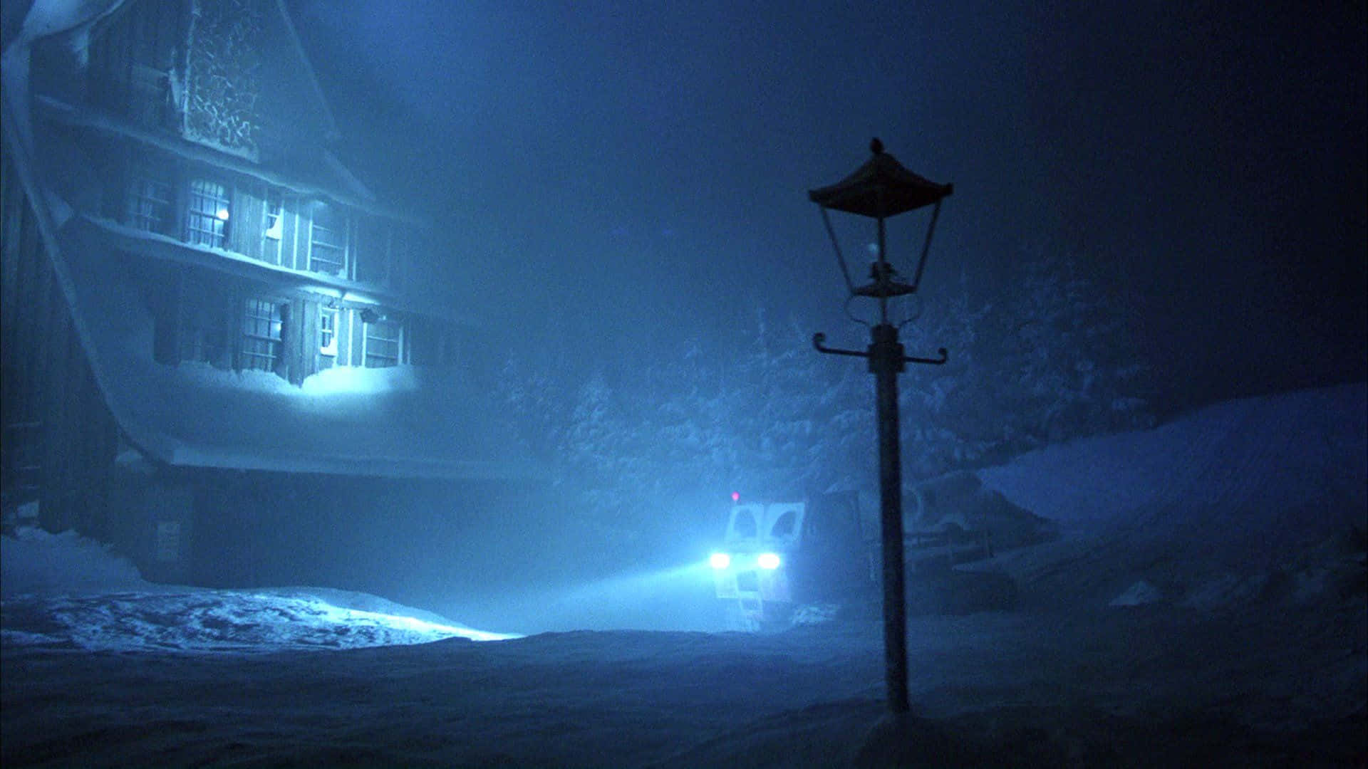  壁紙 — 一間被雪覆蓋的房子在夜晚溫暖地發光，車輛的頭燈穿過霧濛濛的藍色冬日空氣，靠近一盞復古路燈。