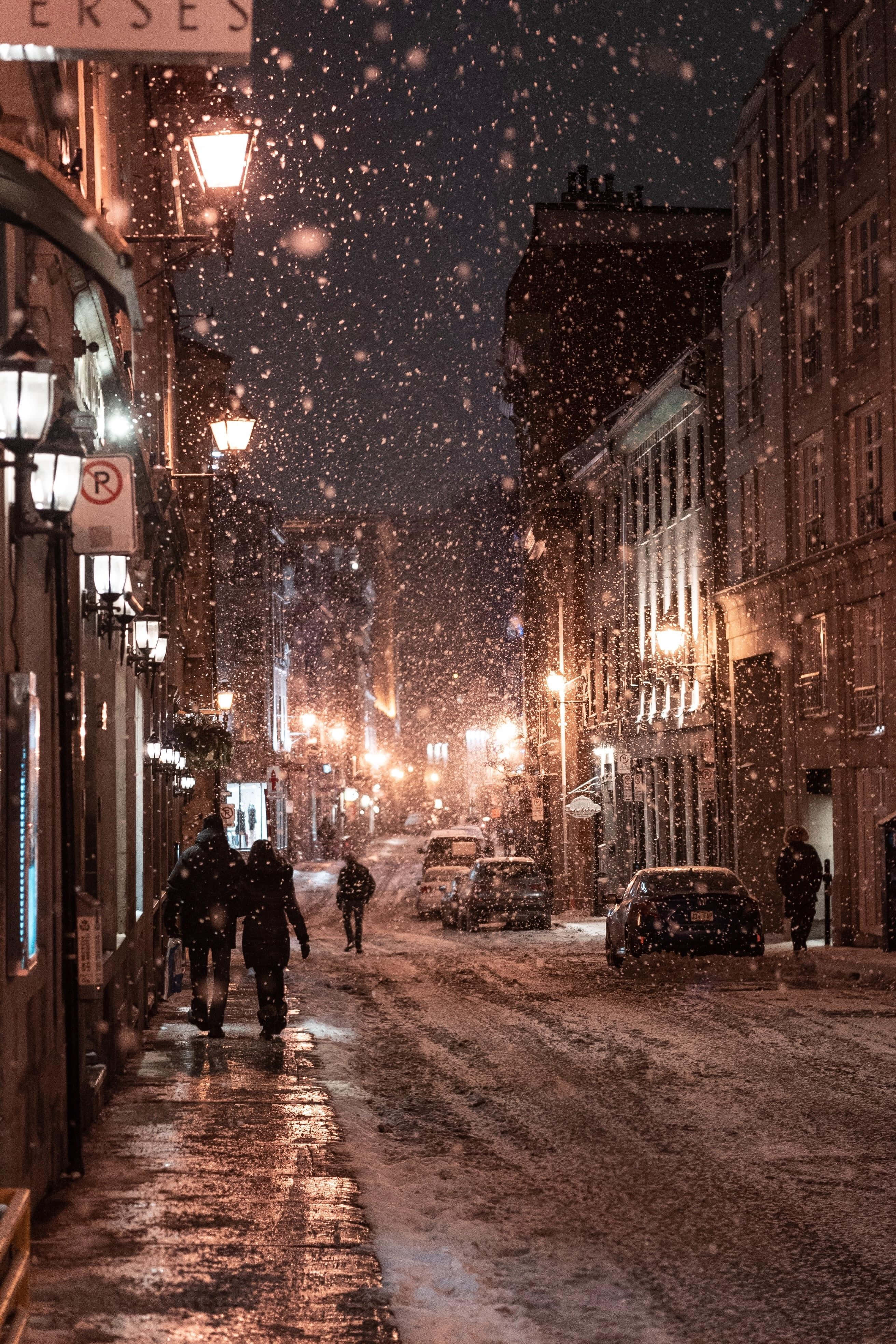 Evening 桌布 壁紙 — 雪花在溫暖的路燈中飄舞，照亮了夜晚寧靜積雪的城市小巷，有行人走過。