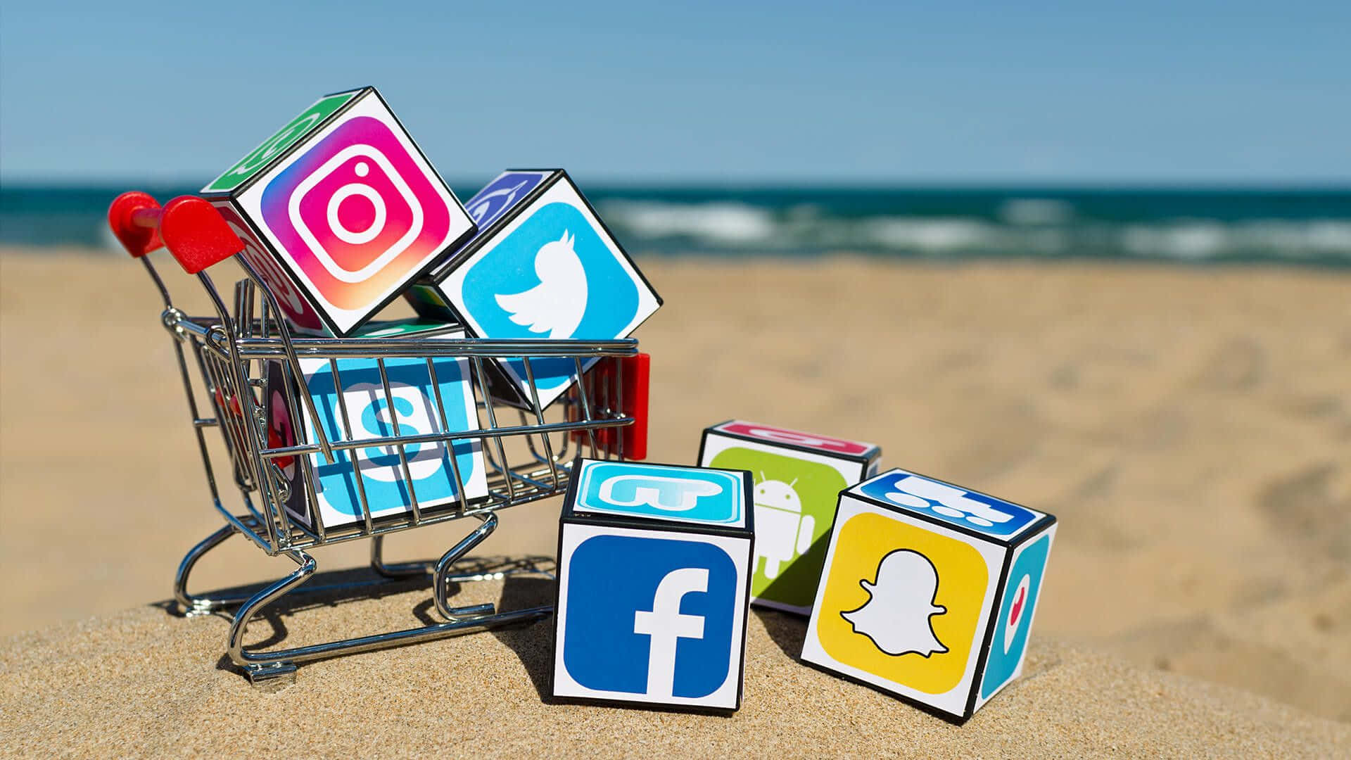 4K Social Media 桌布 壁紙 — 社交媒體應用程式圖示放在彩色立方體上，位於一個微型購物車內，背景是陽光明媚的海灘和海洋波浪。