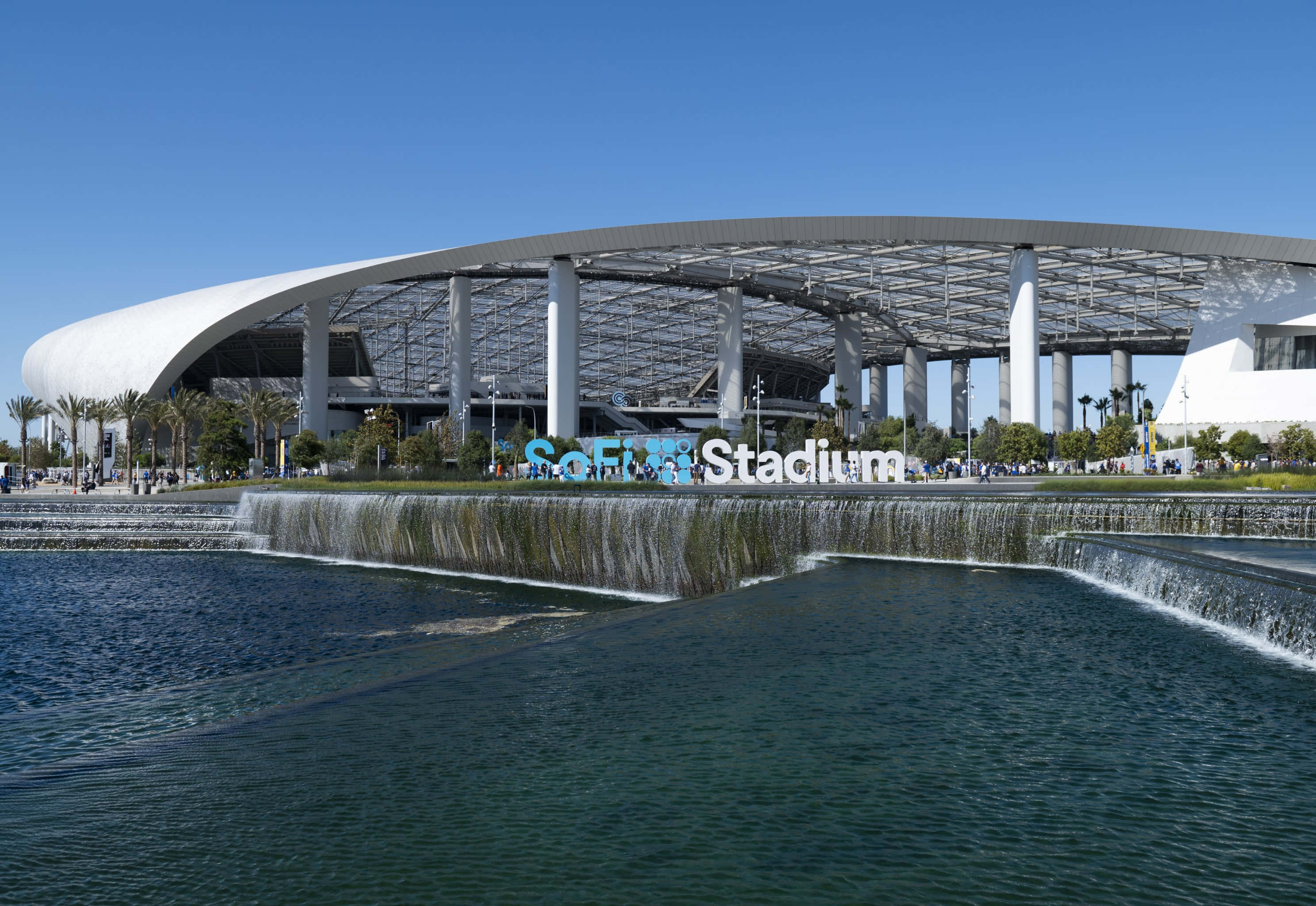 SoFi Stadium重塑體育體驗 壁紙 — 一個現代化的體育場，擁有彎曲的白色屋頂和層疊的水景，在晴朗的藍天下。