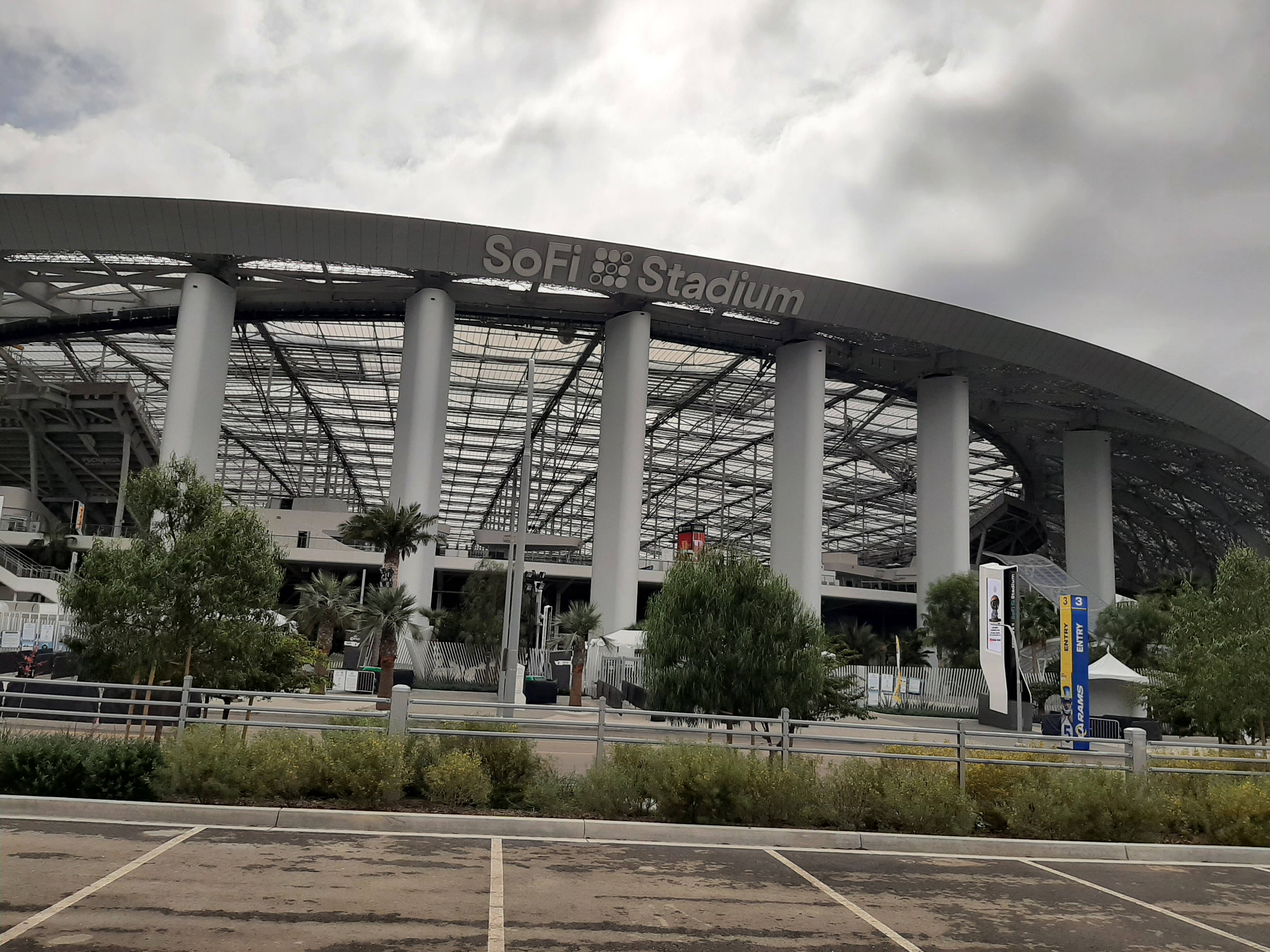 SoFi體育場NFL美式足球賽場 壁紙 — Sofi Stadium 矗立於多雲的天空下，前景有棕櫚樹和停車場。