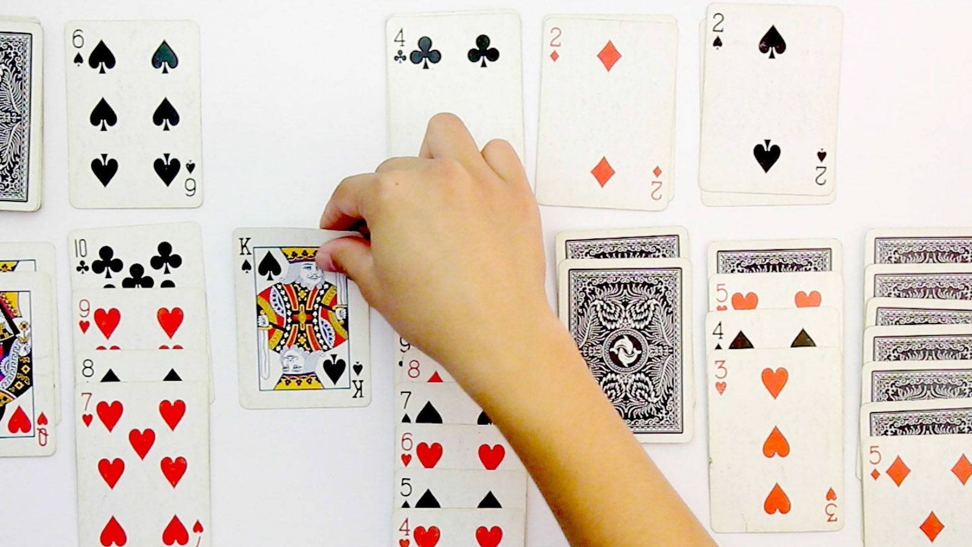 Solitaire 撲克牌：放置國王 壁紙 — 一隻手小心地將一張黑桃K牌放在白色平面上散落的紙牌中。