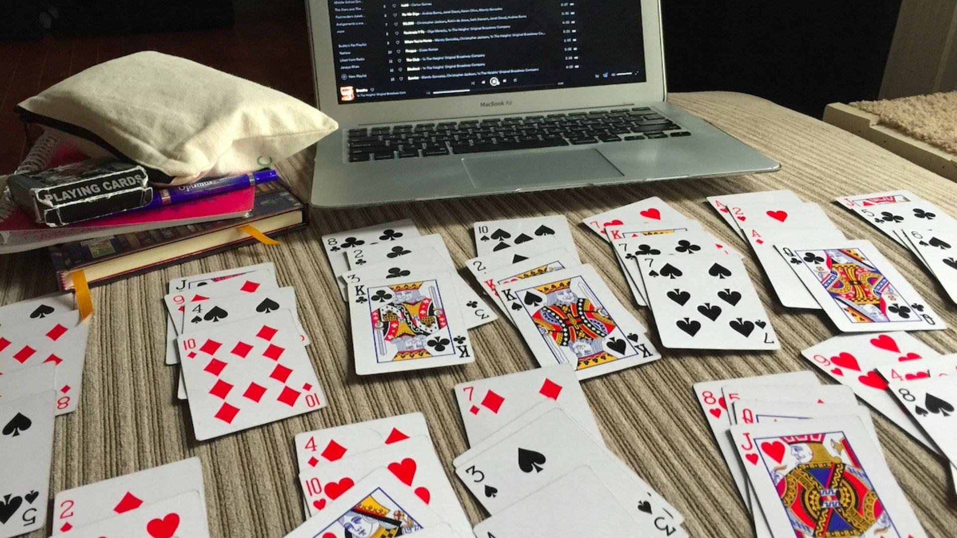 Solitaire 桌上學習 壁紙 — 散落的啤牌和手提電腦放在一張木桌上，周圍有書本和一個枕頭。