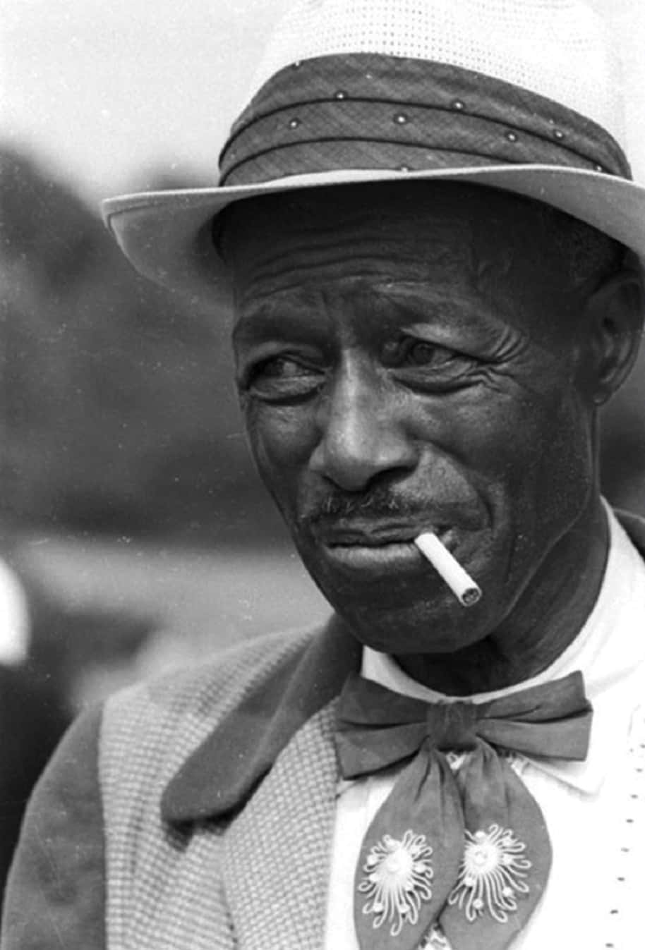 傳奇藍調音樂家 Son House 畫像 壁紙 — 一位戴帽子和領結、嘴裡叼著香煙的長者的黑白肖像。