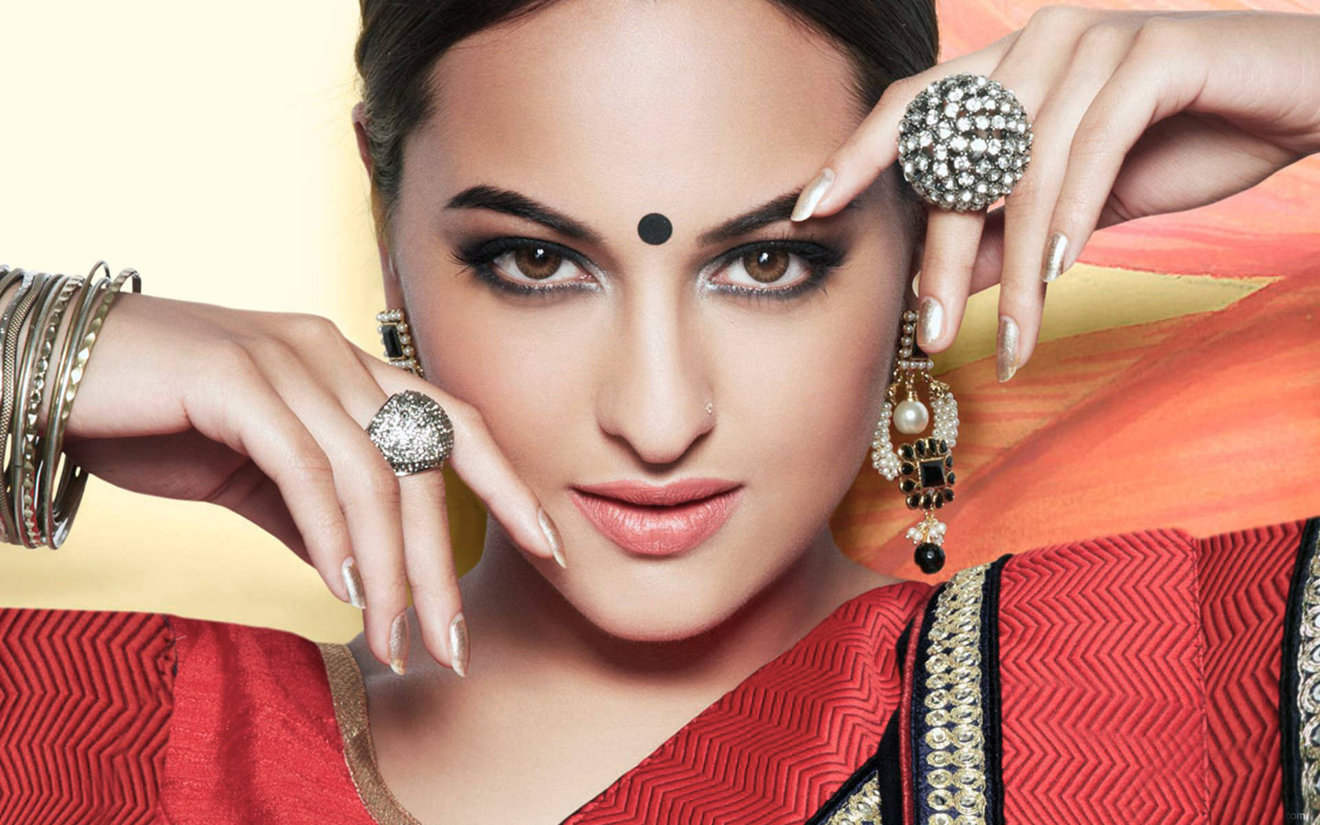 Sonakshi Sinha 的優雅印度風造型 壁紙 — 一位身穿紅色圖案紗麗、飾以精緻珠寶的女性，在溫暖漸變背景前自信地擺姿勢。