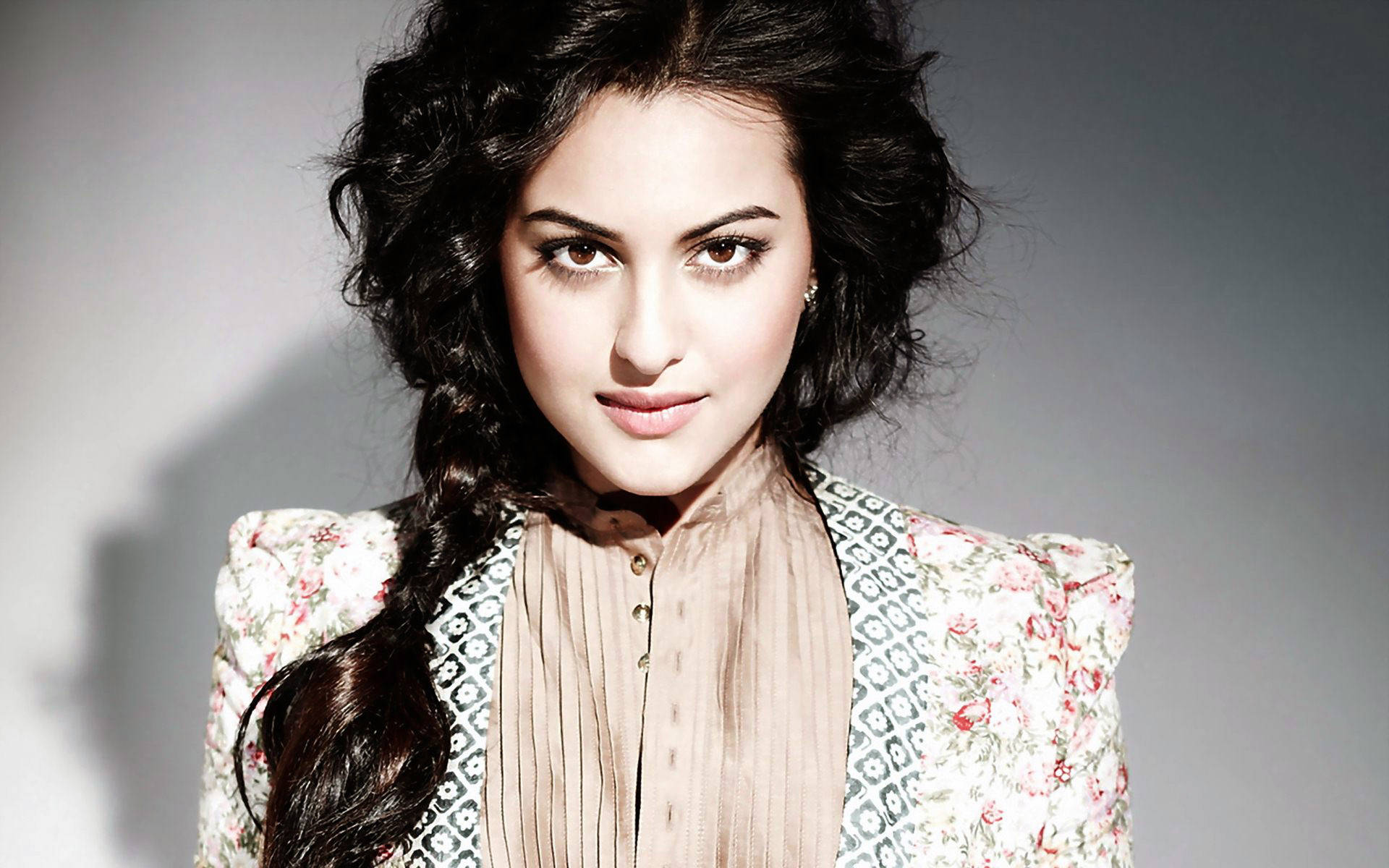 Sonakshi Sinha 優雅造型與風格 壁紙 — 一位擁有深色捲髮和花卉外套的女性，在柔和的灰色工作室背景下自信地凝視。