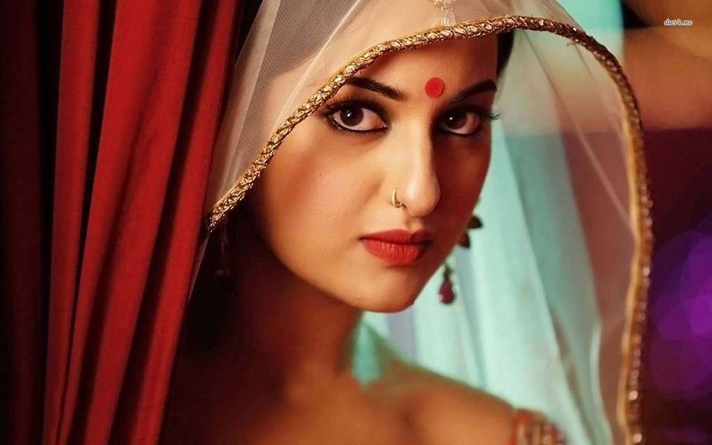 Sonakshi Sinha 印度模特兒紅點造型 壁紙 — 一位身穿傳統印度新娘服飾、額頭點有紅色吉祥痣、戴著金線刺繡頭紗的女子，從華麗的紅色窗簾後探出頭來。