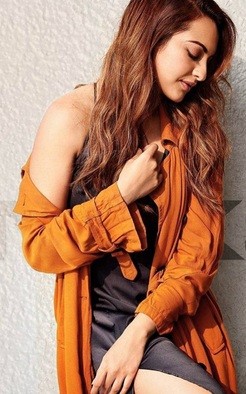 Sonakshi Sinha 情緒化姿勢 壁紙 — 一位長捲髮的女性穿著橙色開襟襯衫，靠在有紋理的白色牆壁上，低頭露出柔和的表情。