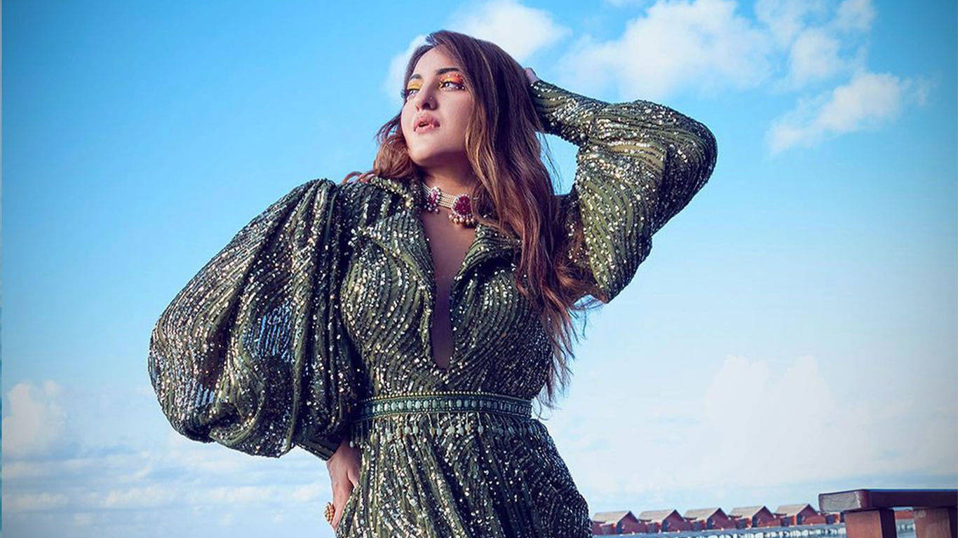 Sonakshi Sinha 時尚綠色洋裝 壁紙 — 一位身穿閃亮綠色連衣裙的女性在藍天白雲下自信地擺姿勢。