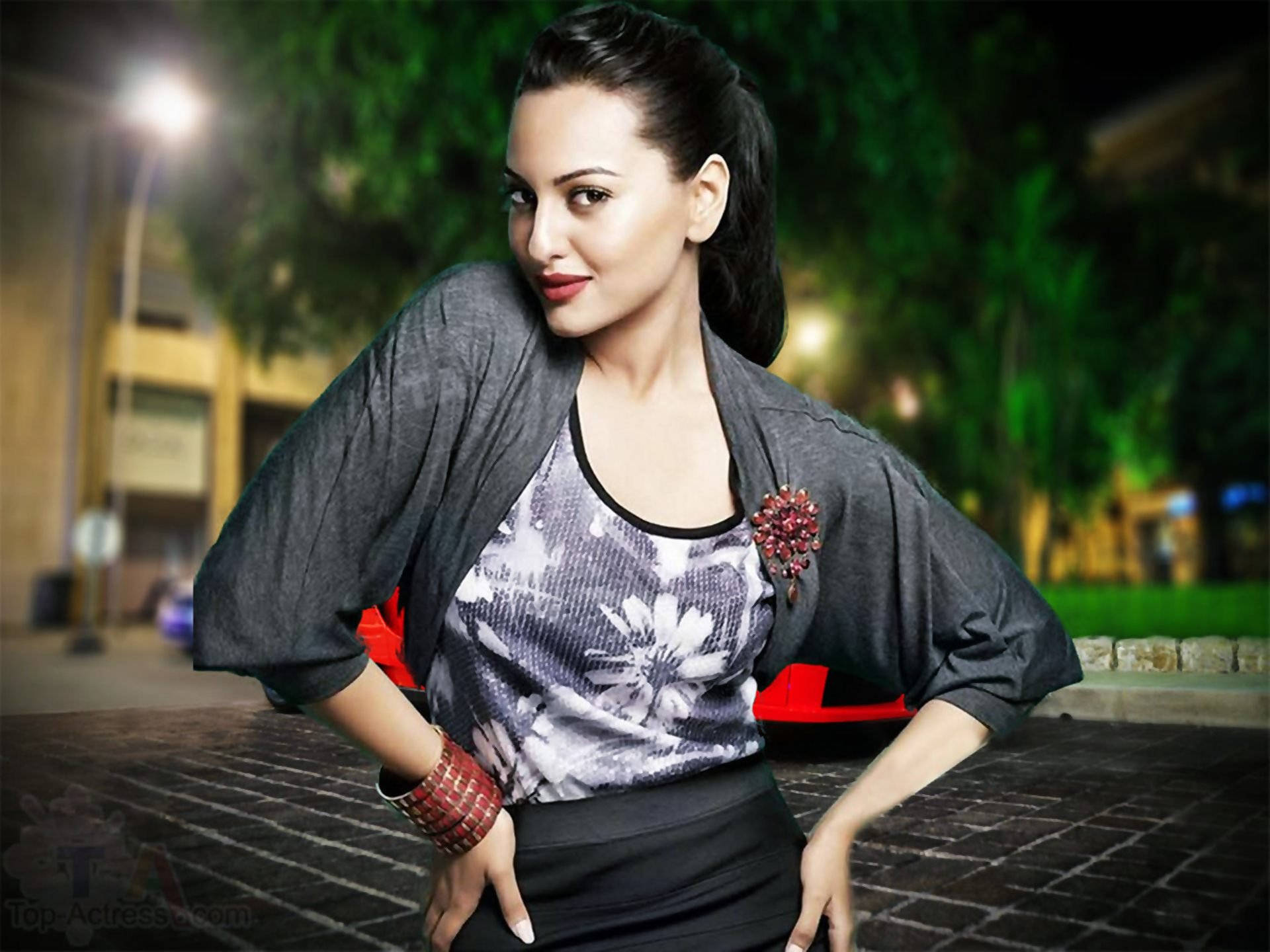 Sonakshi Sinha 的高雅造型 壁紙 — 一位身穿灰色開襟衫和花卉上衣的女性，在夜晚的路燈下自信地站在磚砌人行道上擺姿勢。