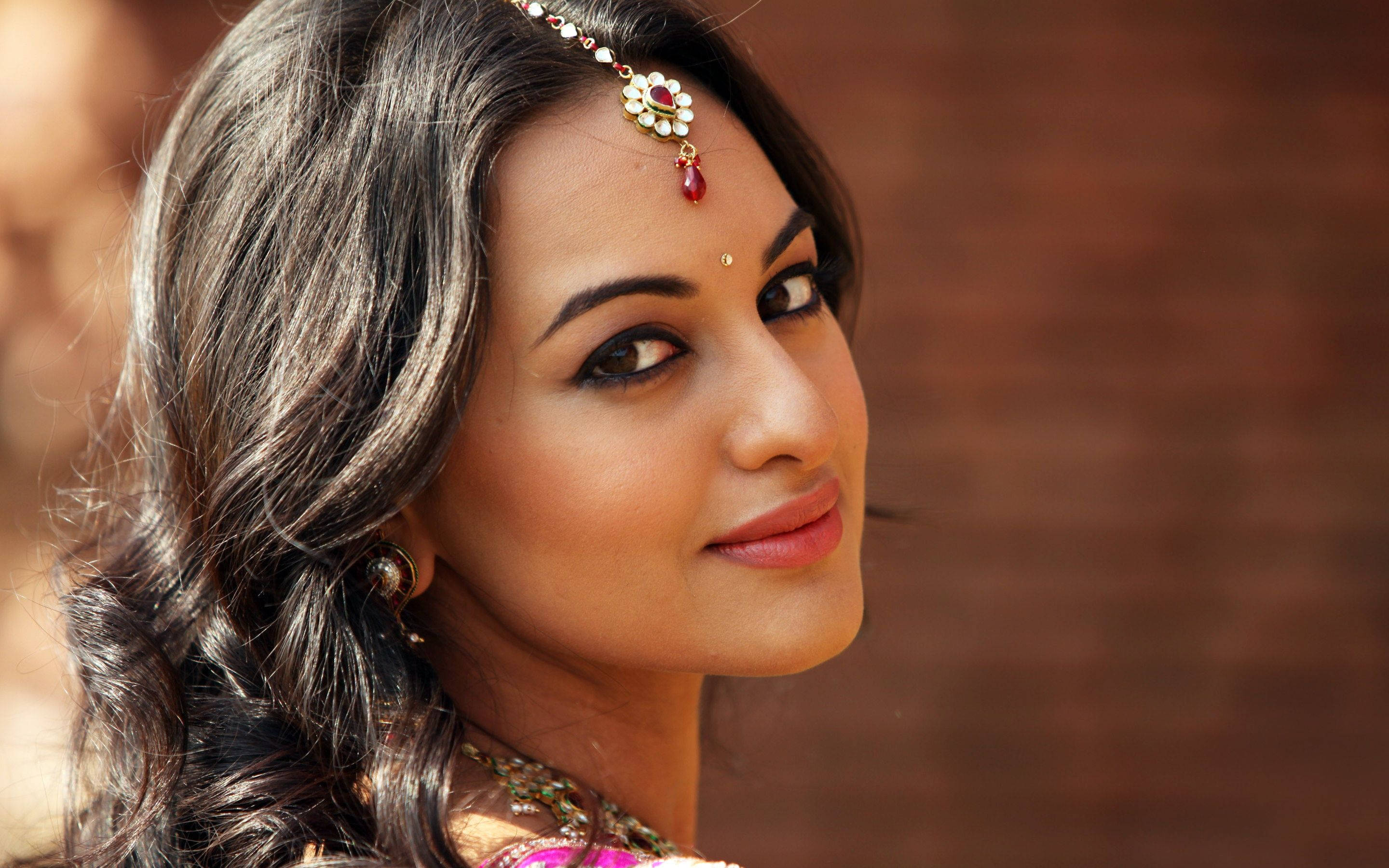 Sonakshi Sinha 精緻妝容 壁紙 — 一位容光煥發的女性，深色波浪髮上飾有紅色寶石Maang Tikka，在溫暖模糊的磚牆背景下溫柔微笑。